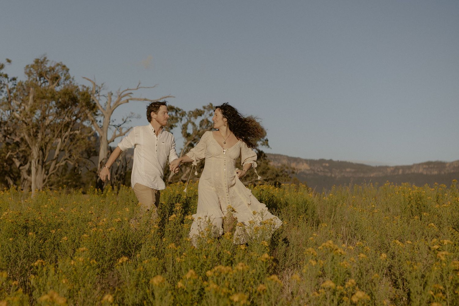 Romantic couple embracing in a sunlit wildflower field with mountains in the background.
