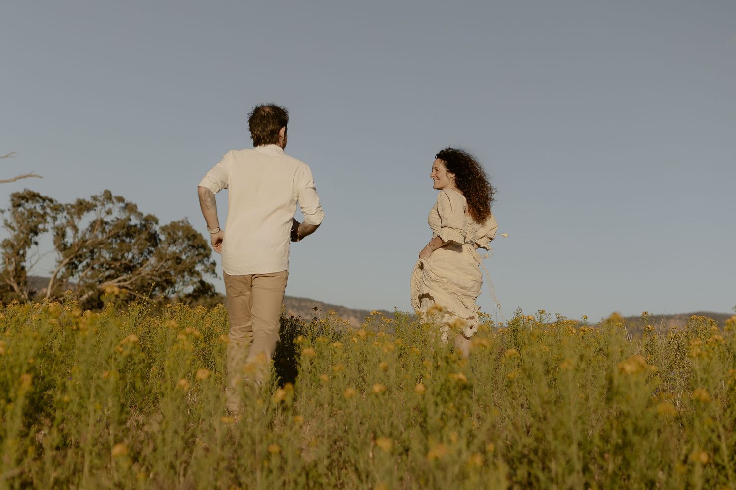 Two figures walk together through a sunlit meadow of tall grass and wildflowers.