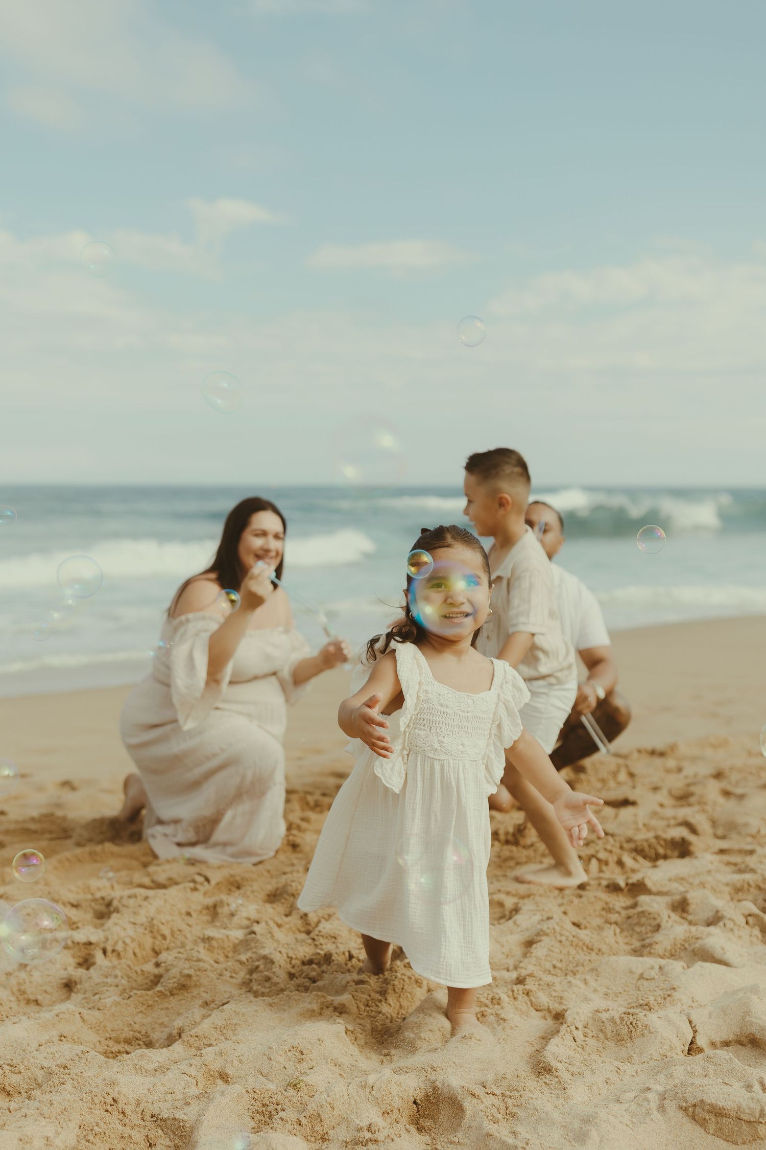 Family members in white clothing interact playfully on a sunny beach with waves and blue sky.
