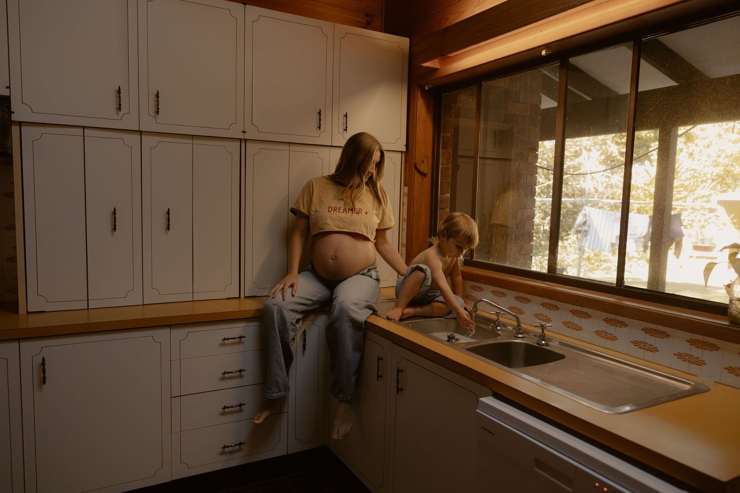 Pregnant mother sits on kitchen counter in cozy wood-paneled kitchen while young child plays at sink.