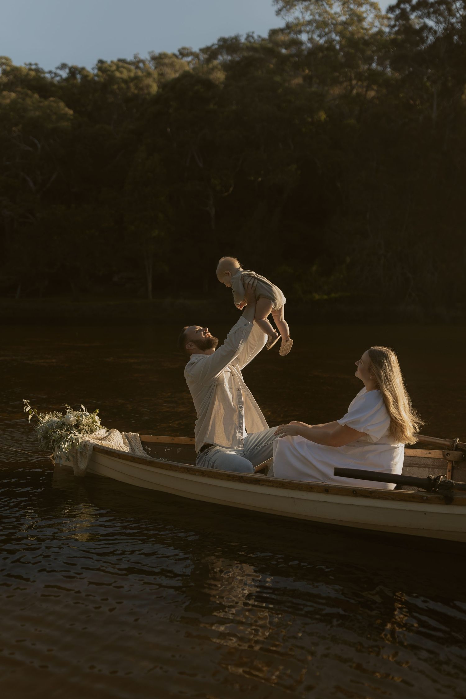 A couple in white outfits lifts their baby in a flower-decorated boat on a tranquil lake during golden hour.