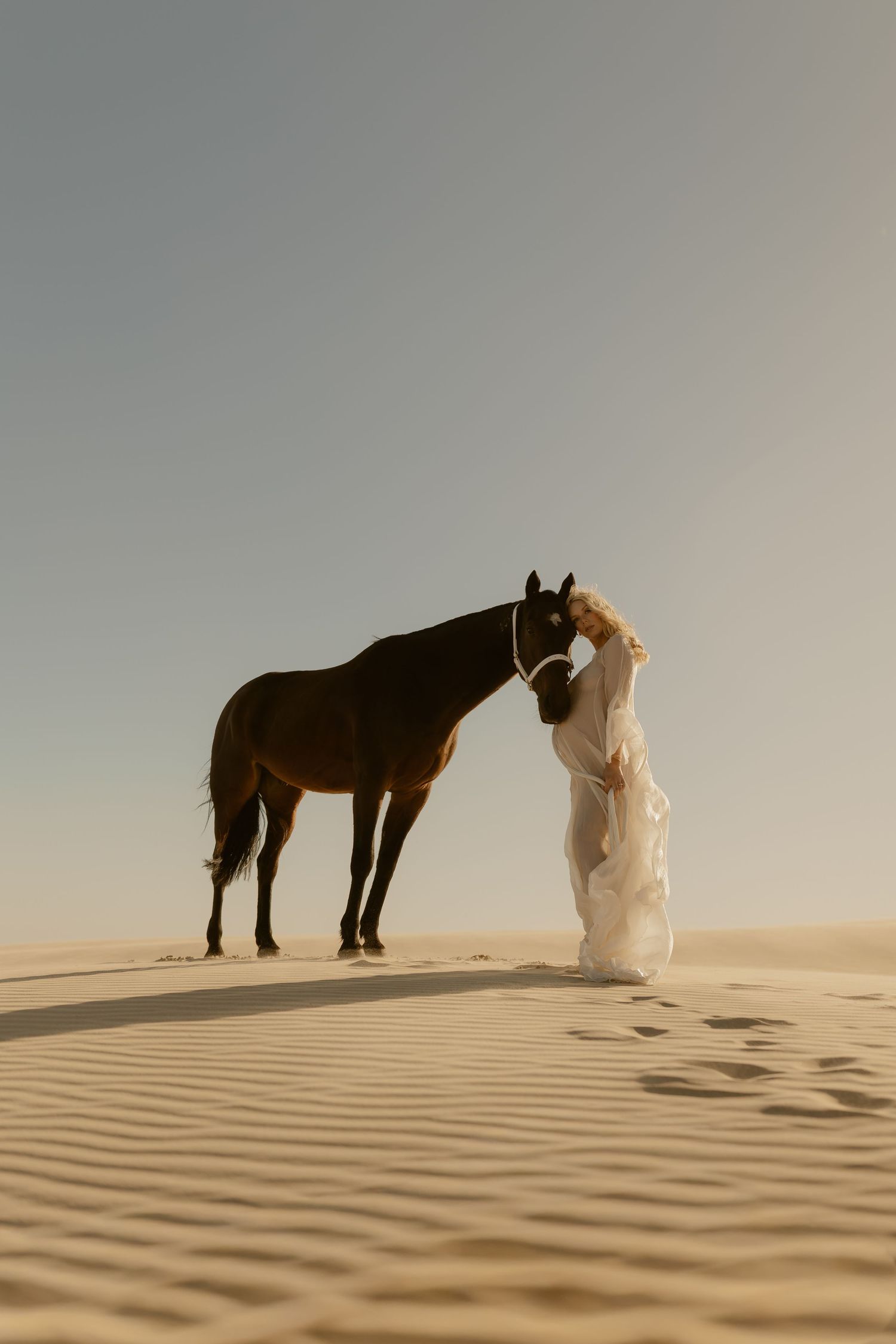 A bride in a white gown stands with a dark horse on golden sand dunes in a serene desert landscape.