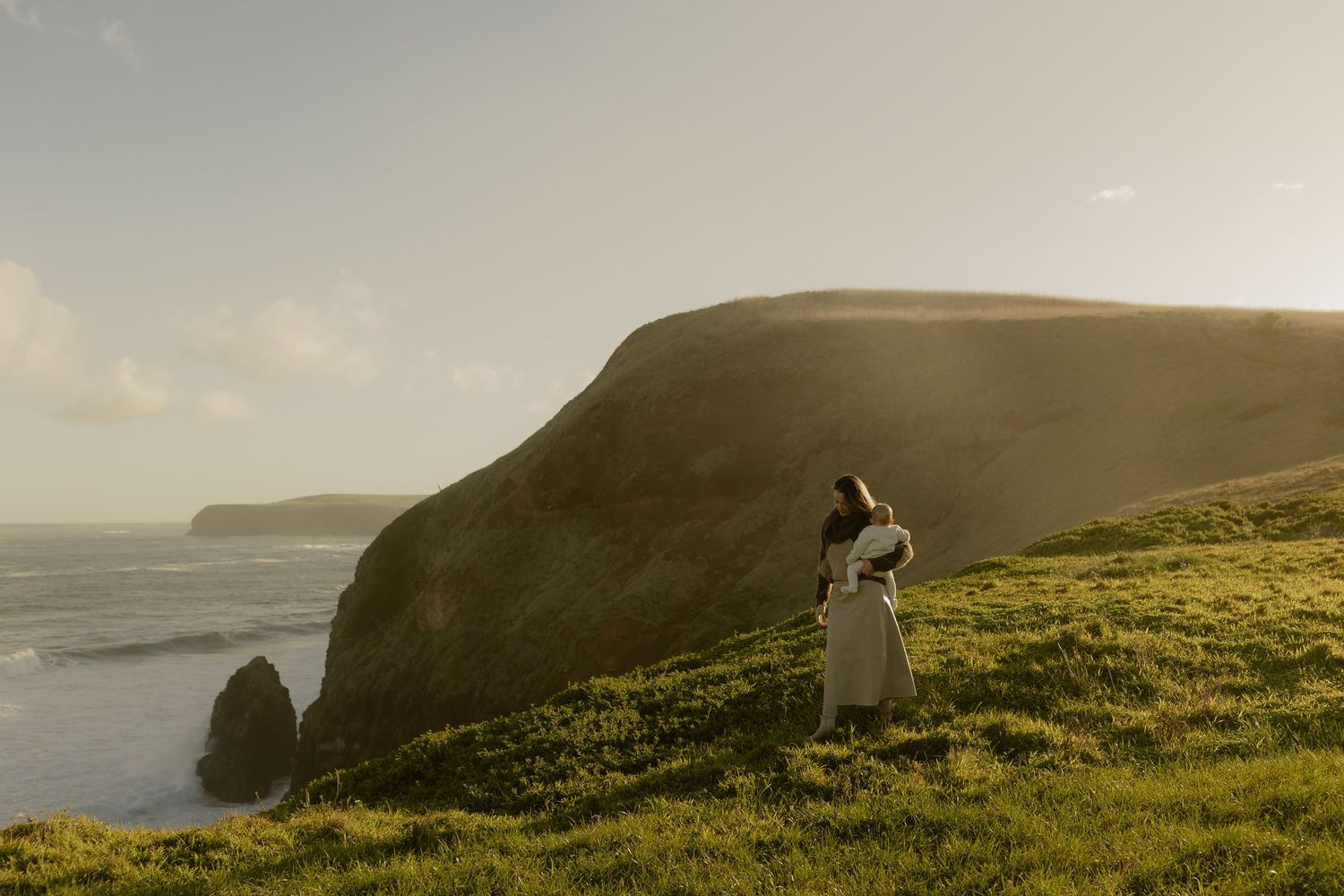Woman in white dress standing on green coastal cliffs overlooking the ocean at golden hour.