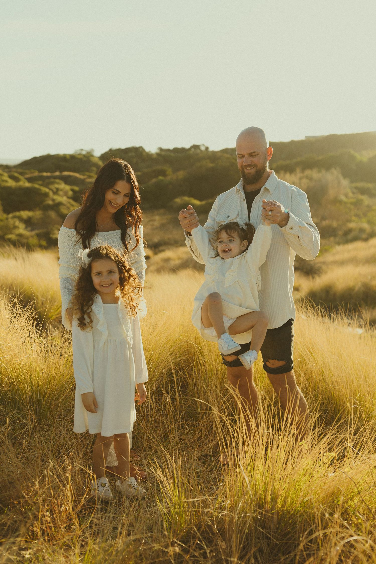 A family wearing white outfits plays together in golden sunlit grass during sunset in a beautiful outdoor setting.