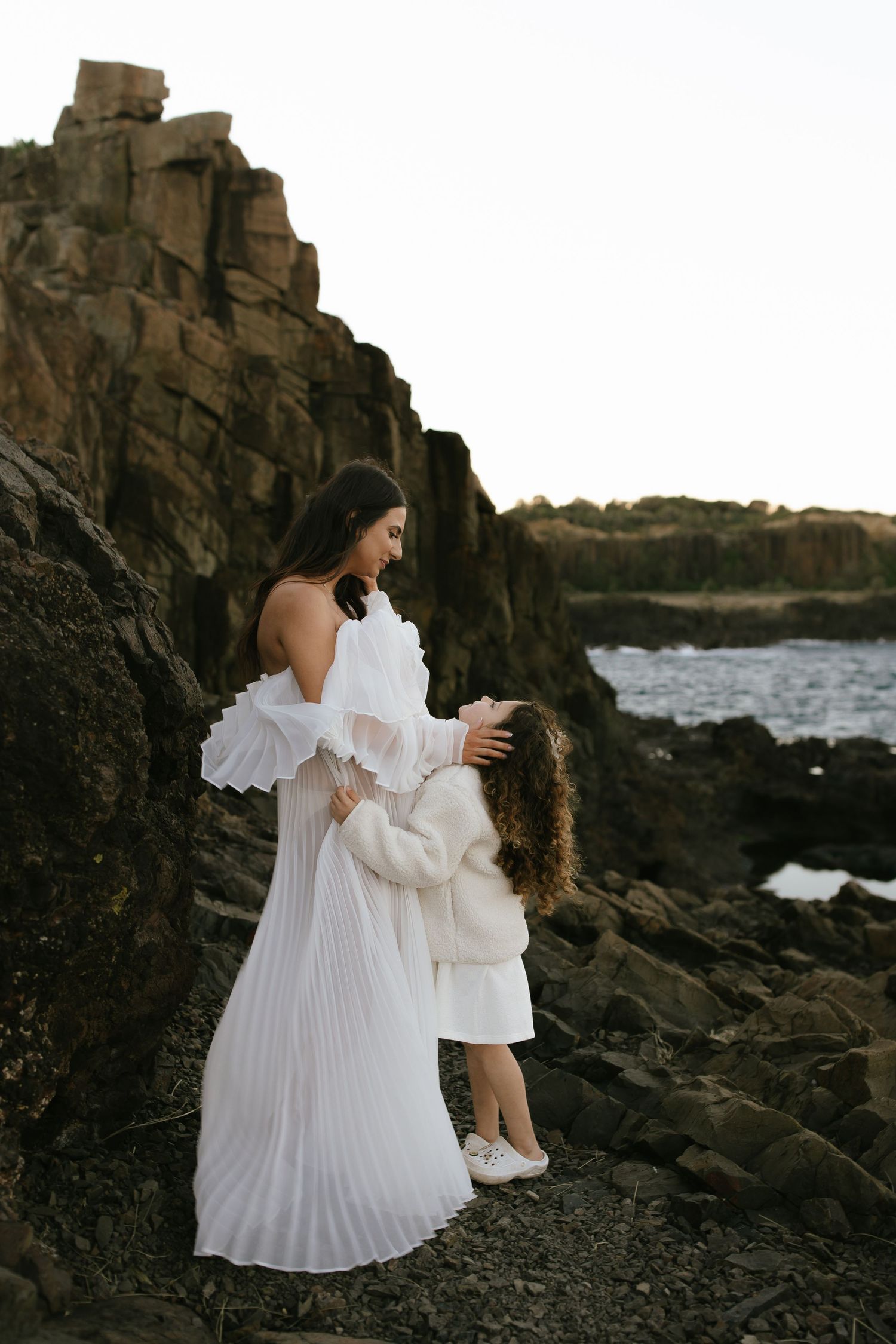 Person wearing ethereal white pleated dress poses among dark volcanic rocks with crashing waves in background.