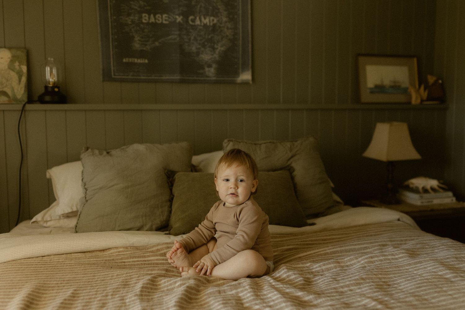 A baby sits on a neutral-toned bed with pillows beneath a wooden wall with framed artwork.
