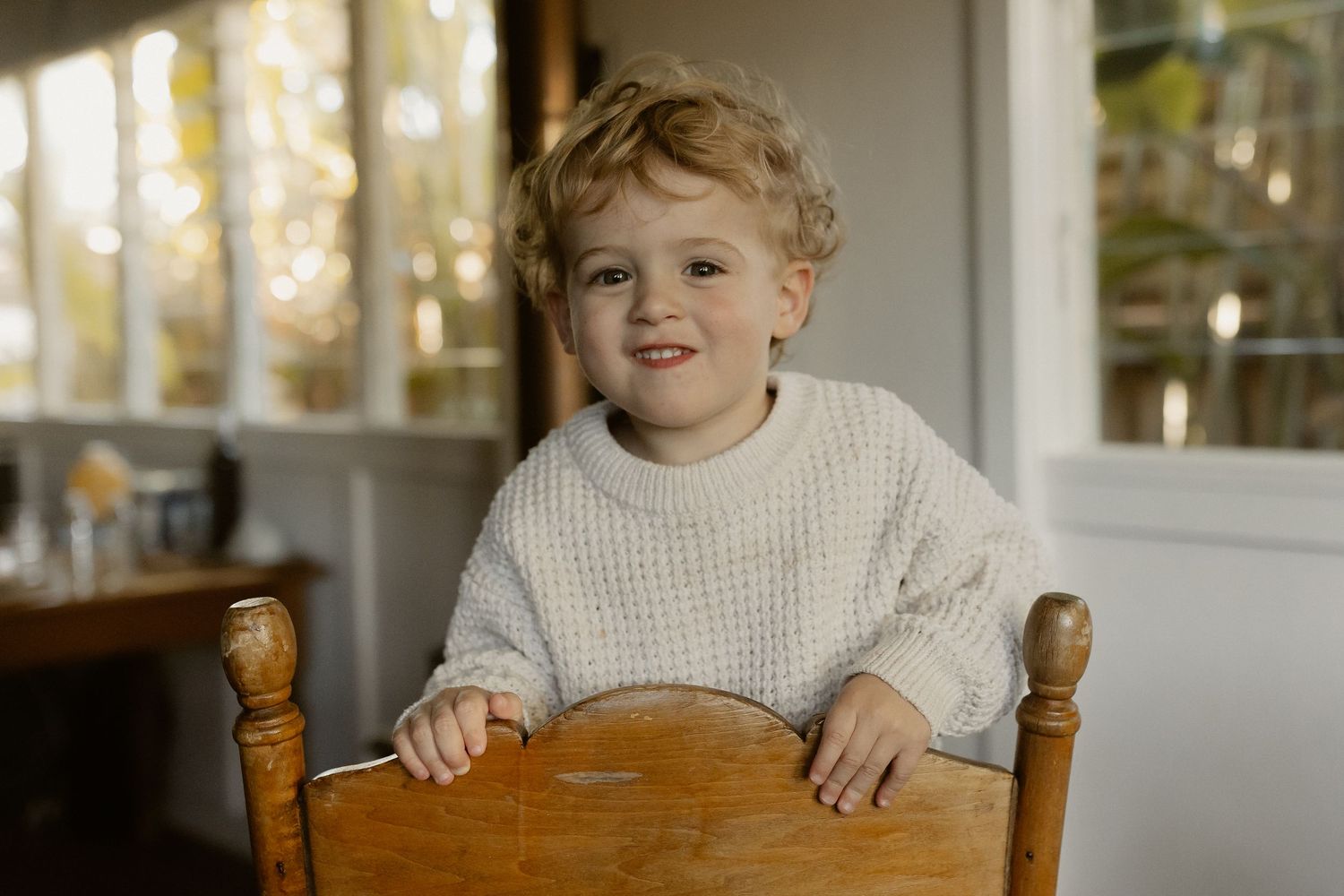 A child in a white sweater sits at a wooden table near bright windows.