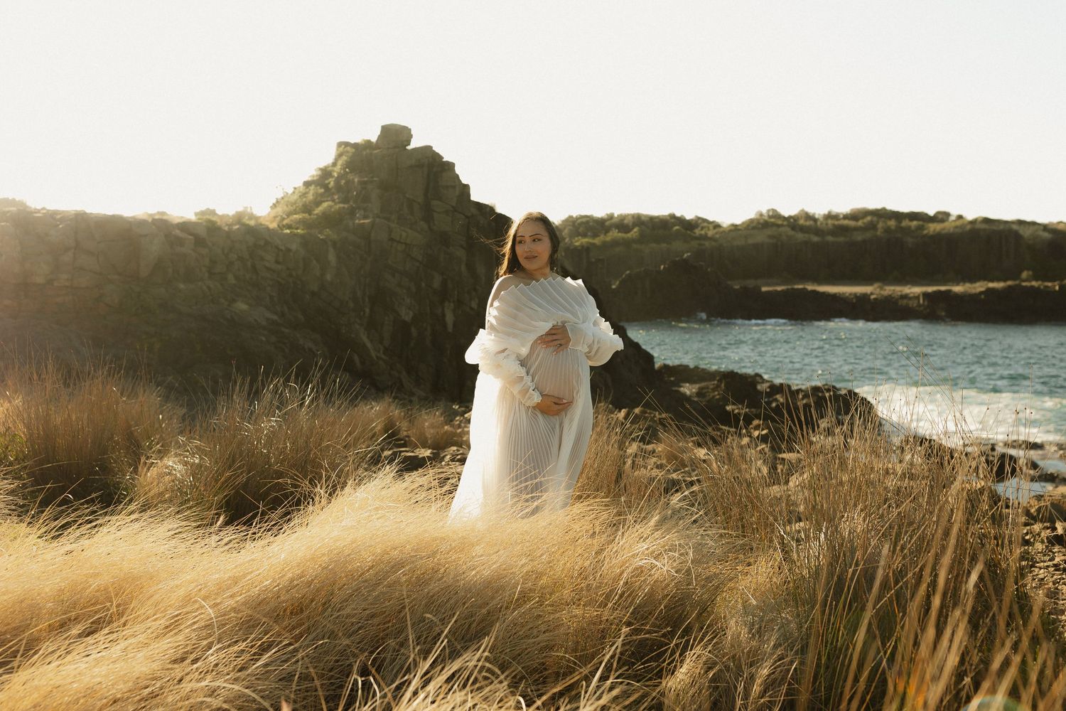 Golden hour maternity photos in a grassy seaside field with sun rays illuminating the natural landscape.