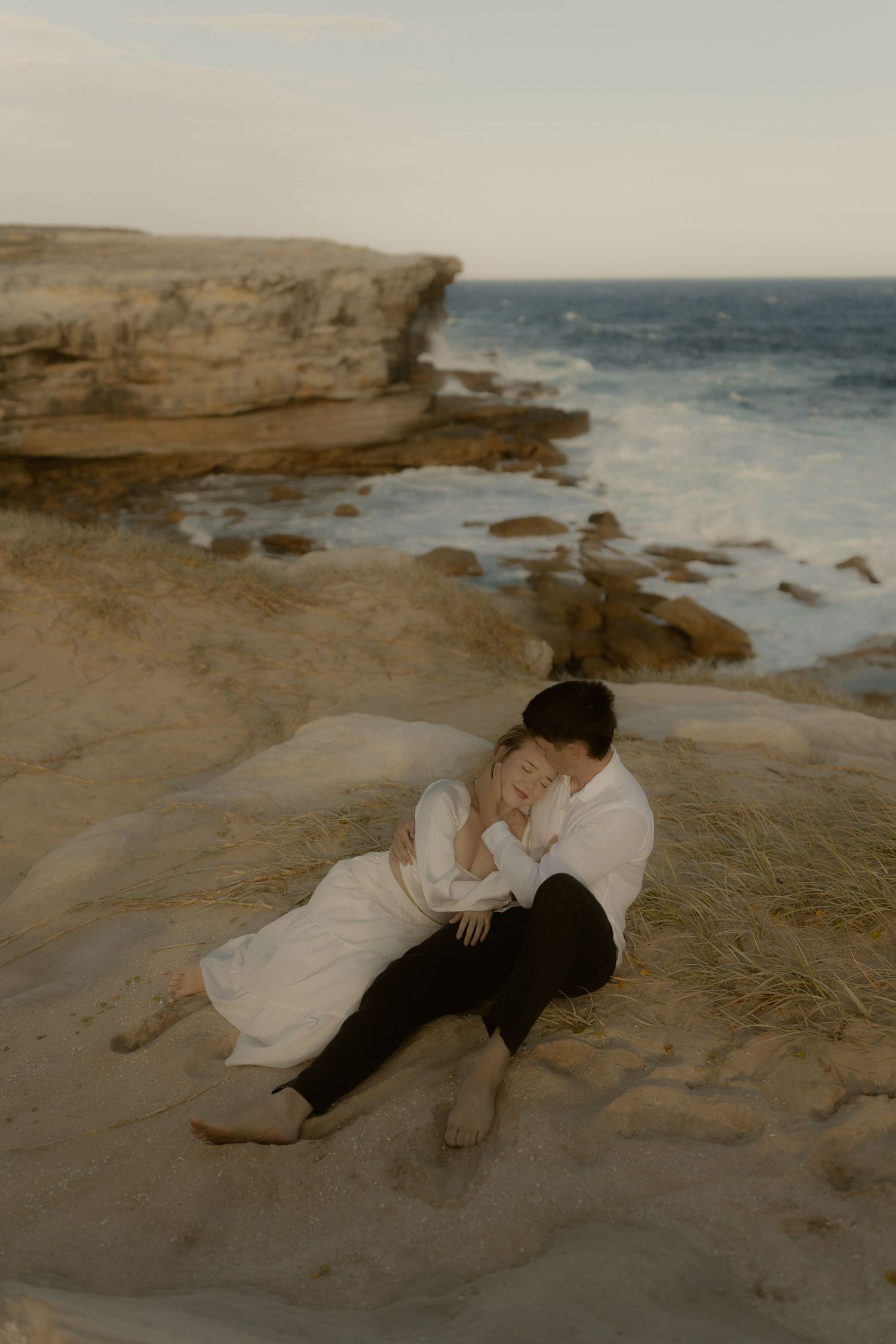 Couple resting together on coastal rocks with ocean waves crashing in background.