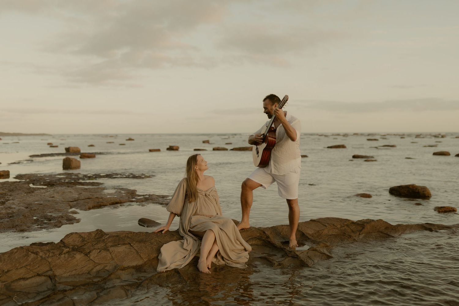 People exploring a rocky beach during sunset while wearing light colored clothing and walking near the ocean's edge.