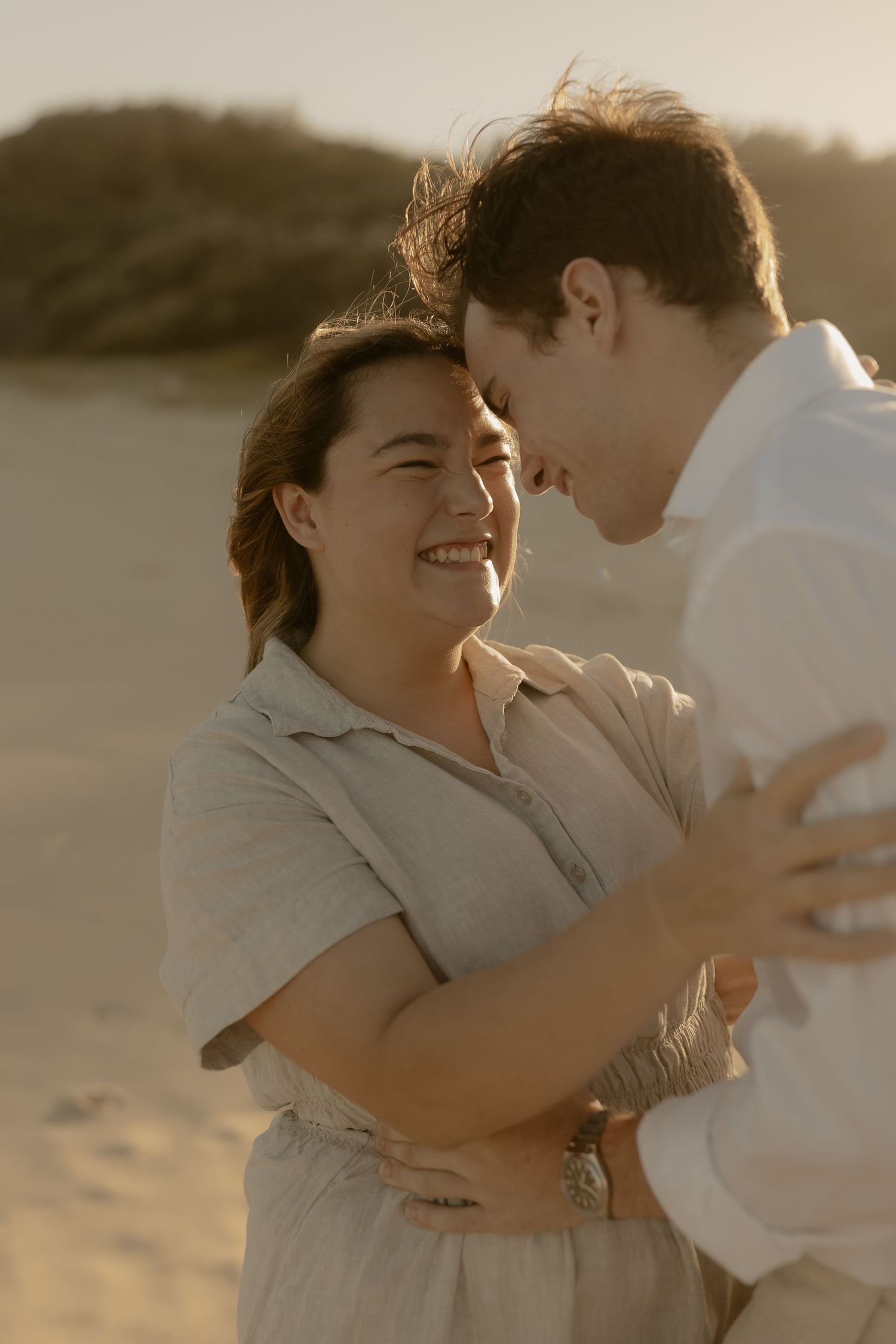 Playful couple laughing and hugging on a sandy beach in warm sunlight.