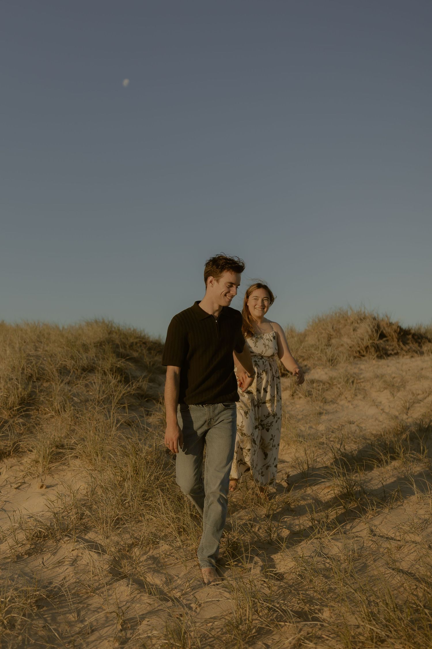 Two people walk together on a sandy hill against a dusky sky in a desert landscape.