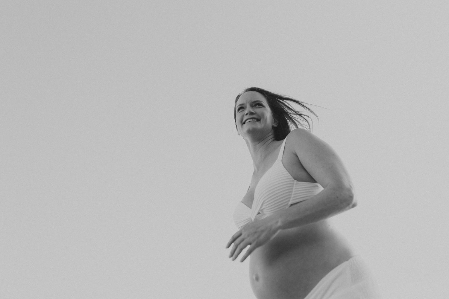 Black and white artistic maternity photo of an expectant mother against a light sky with wind-swept hair.