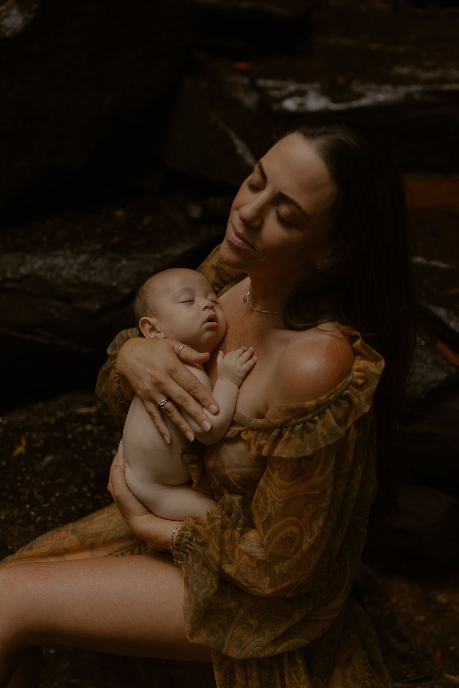 Intimate moody portrait of mother and sleeping infant in vintage-style dress against dark background.