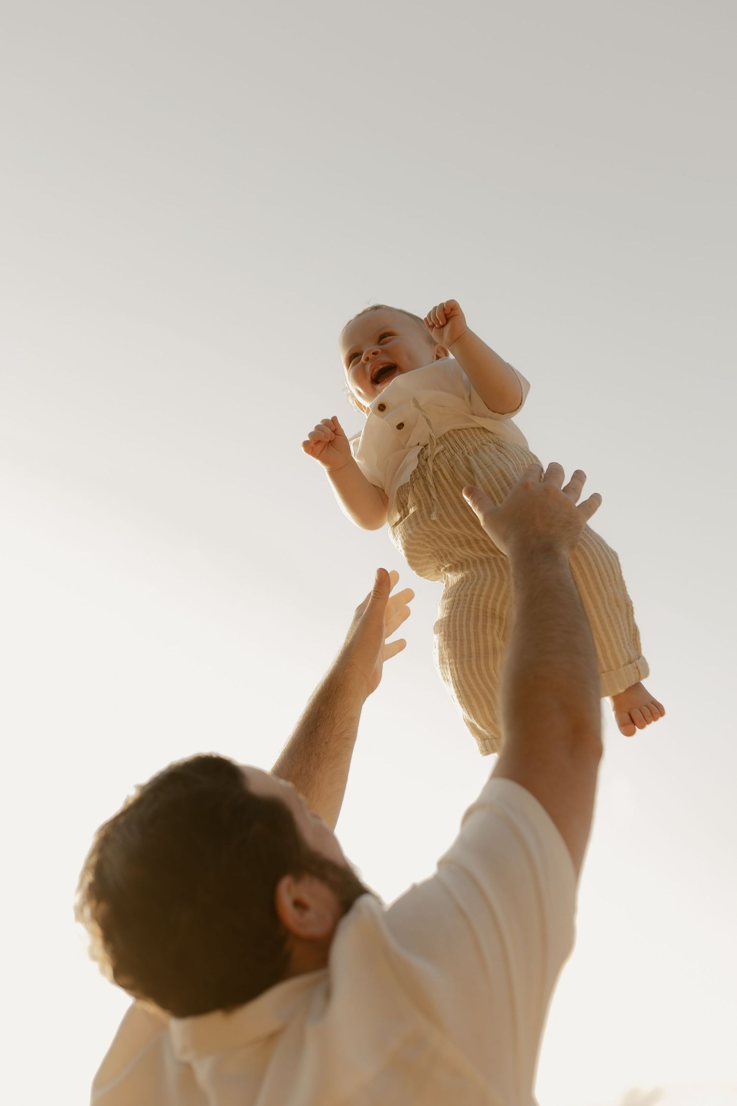 Parent lifts baby up against bright backlit sky in joyful bonding moment.