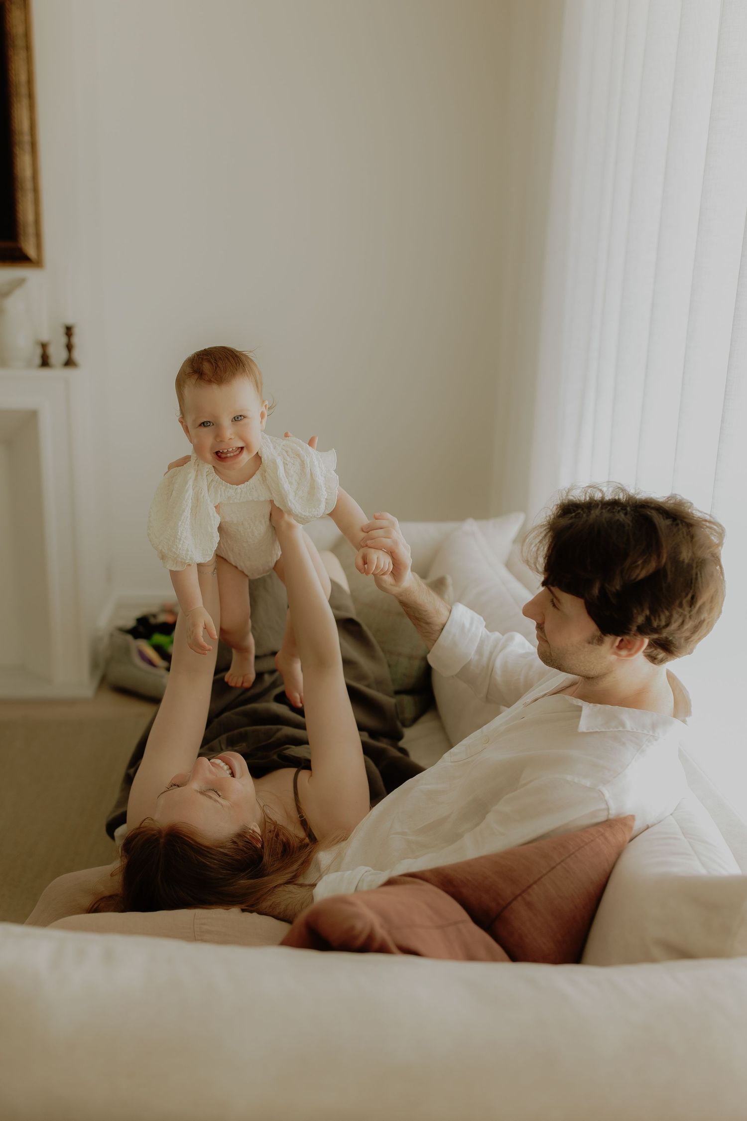A joyful family plays on a white couch, with parents lifting their laughing baby in a bright, cozy living room.