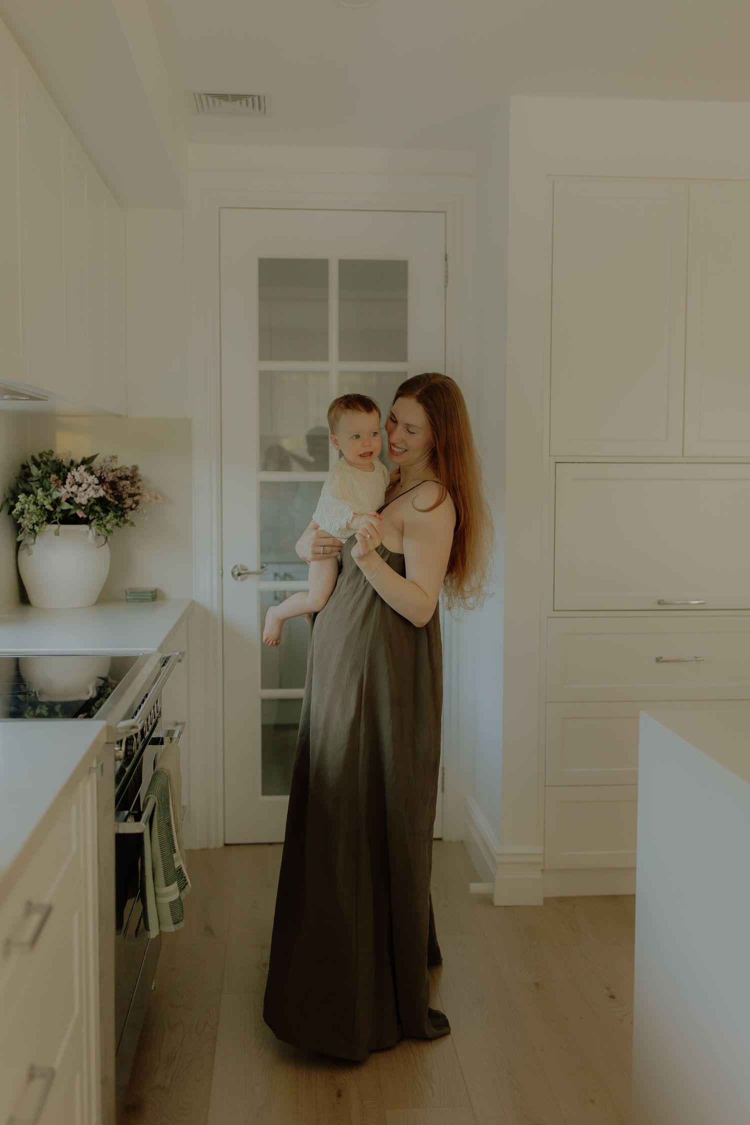 A mother in an olive maxi dress holds her baby in a bright white modern kitchen.