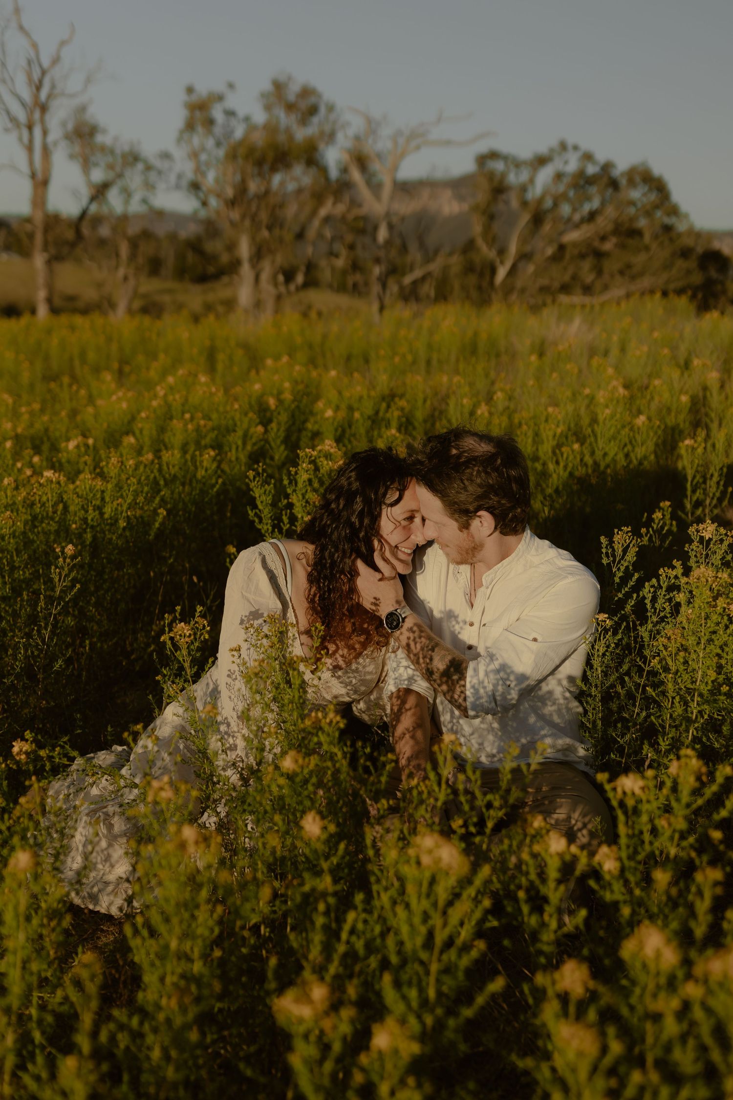 Couple sitting together laughing in a golden wildflower meadow during sunset, sharing a romantic moment.