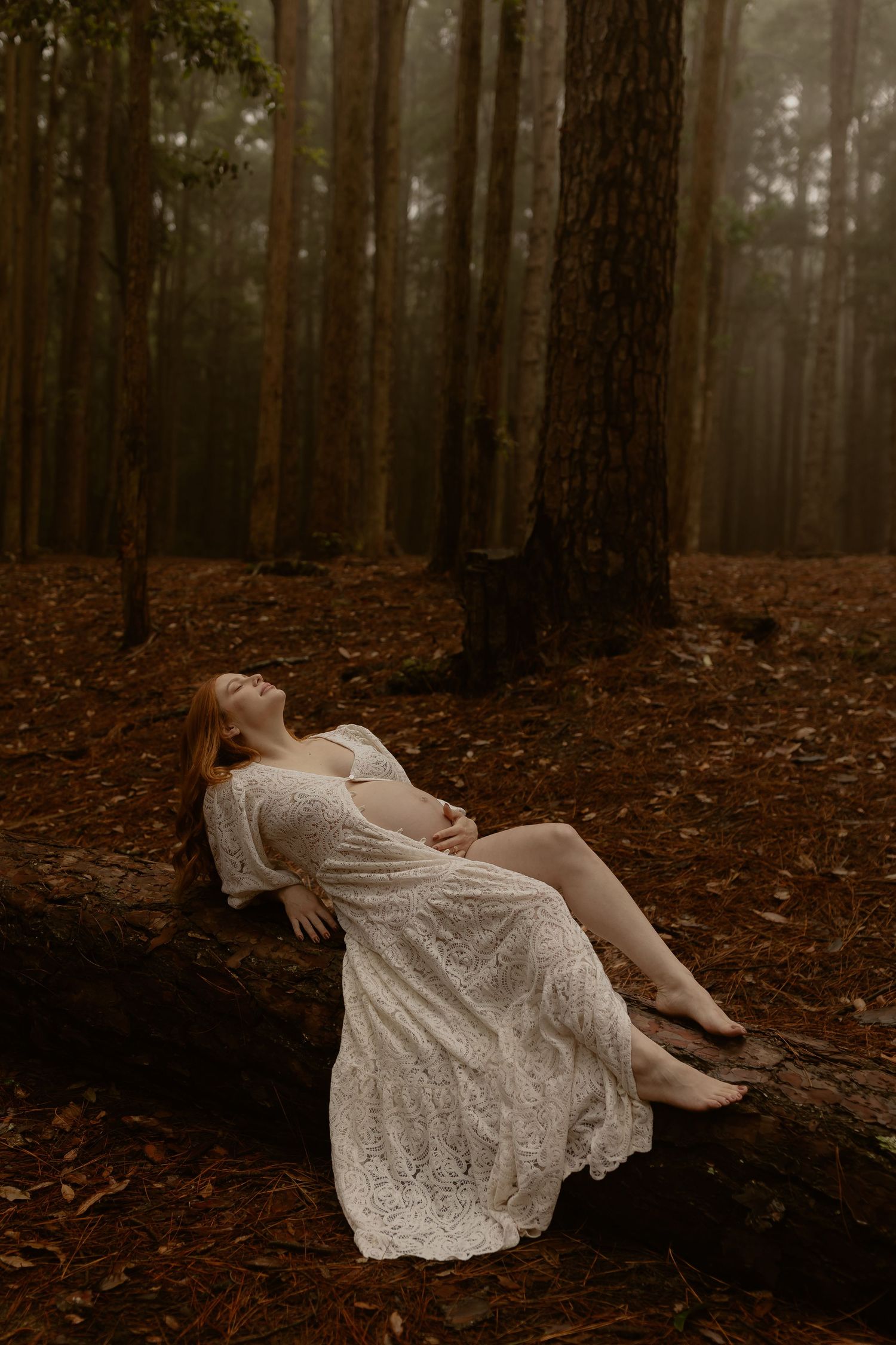 Pregnant woman in white lace dress reclining on log in misty forest.