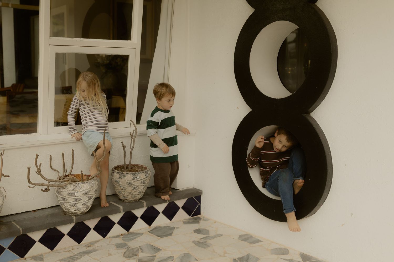 Two children playing on a porch with circular wall decor, one sitting on a windowsill and another inside a round mirror cutout.