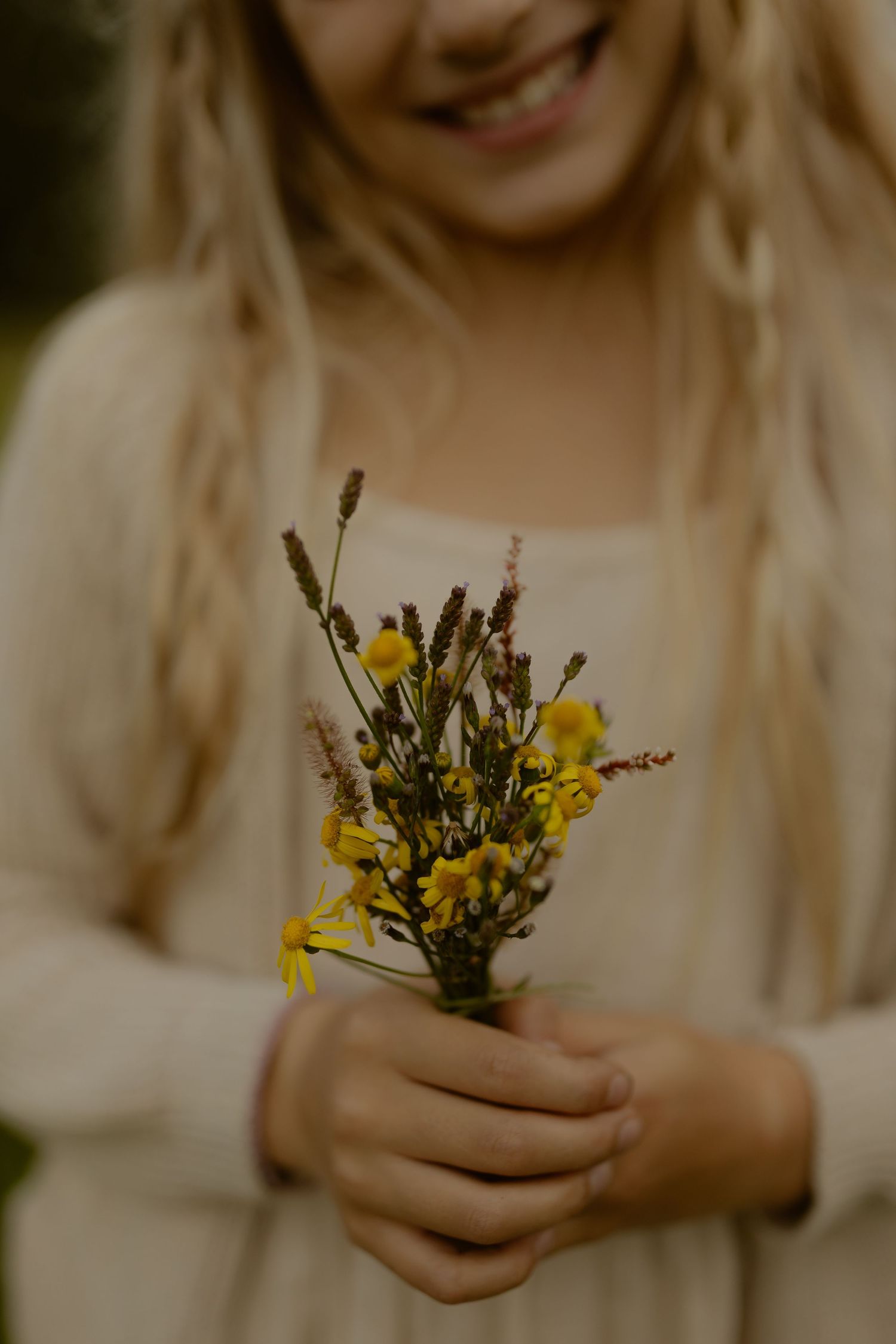 A smiling girl with blonde braids holds a small bouquet of yellow wildflowers.