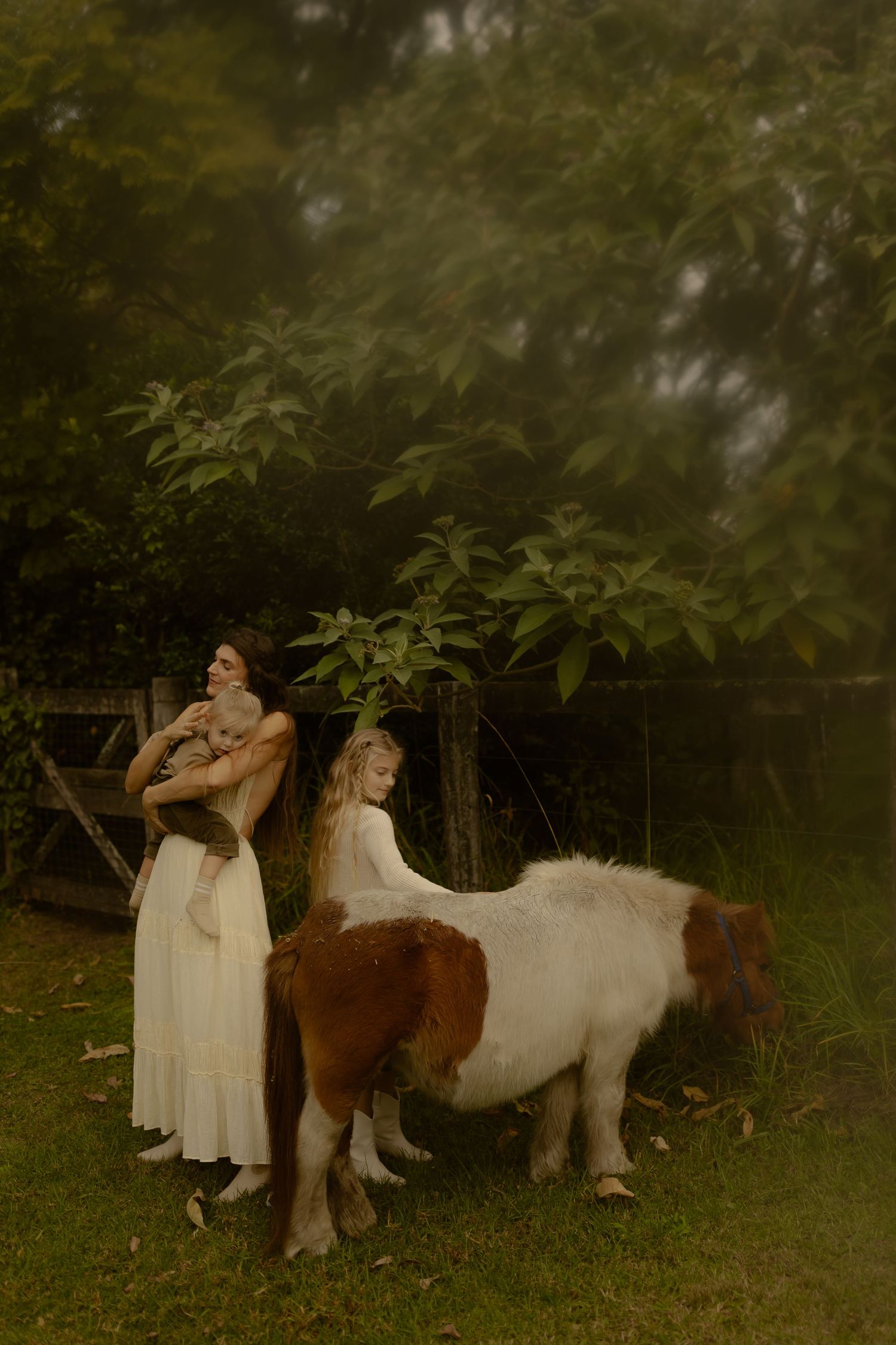 Woman in white dress holding cat beside girl petting a brown and white pony in a lush green garden.