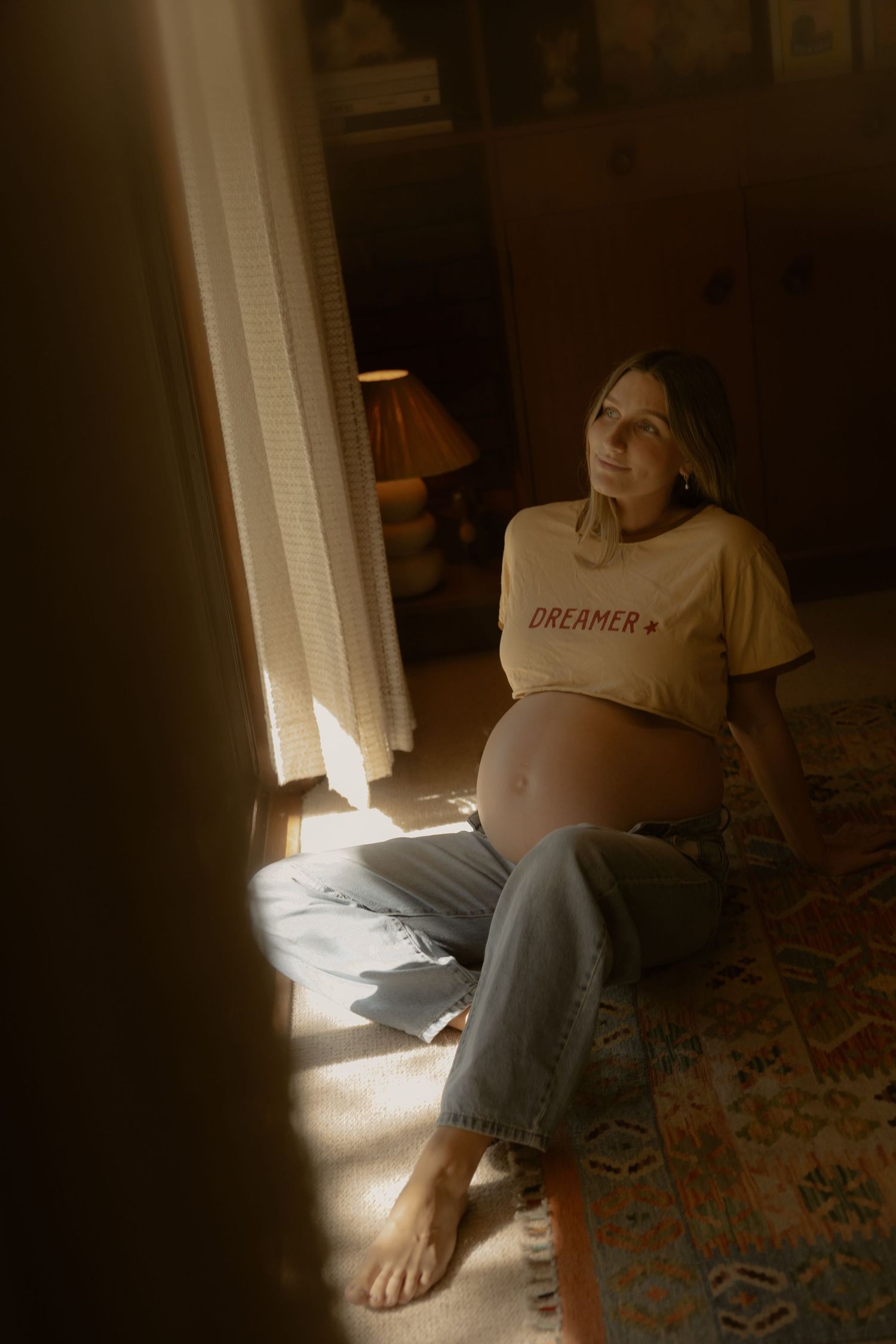 Pregnant woman in a yellow Dreamer crop top and jeans sitting on floor in warm sunlight.