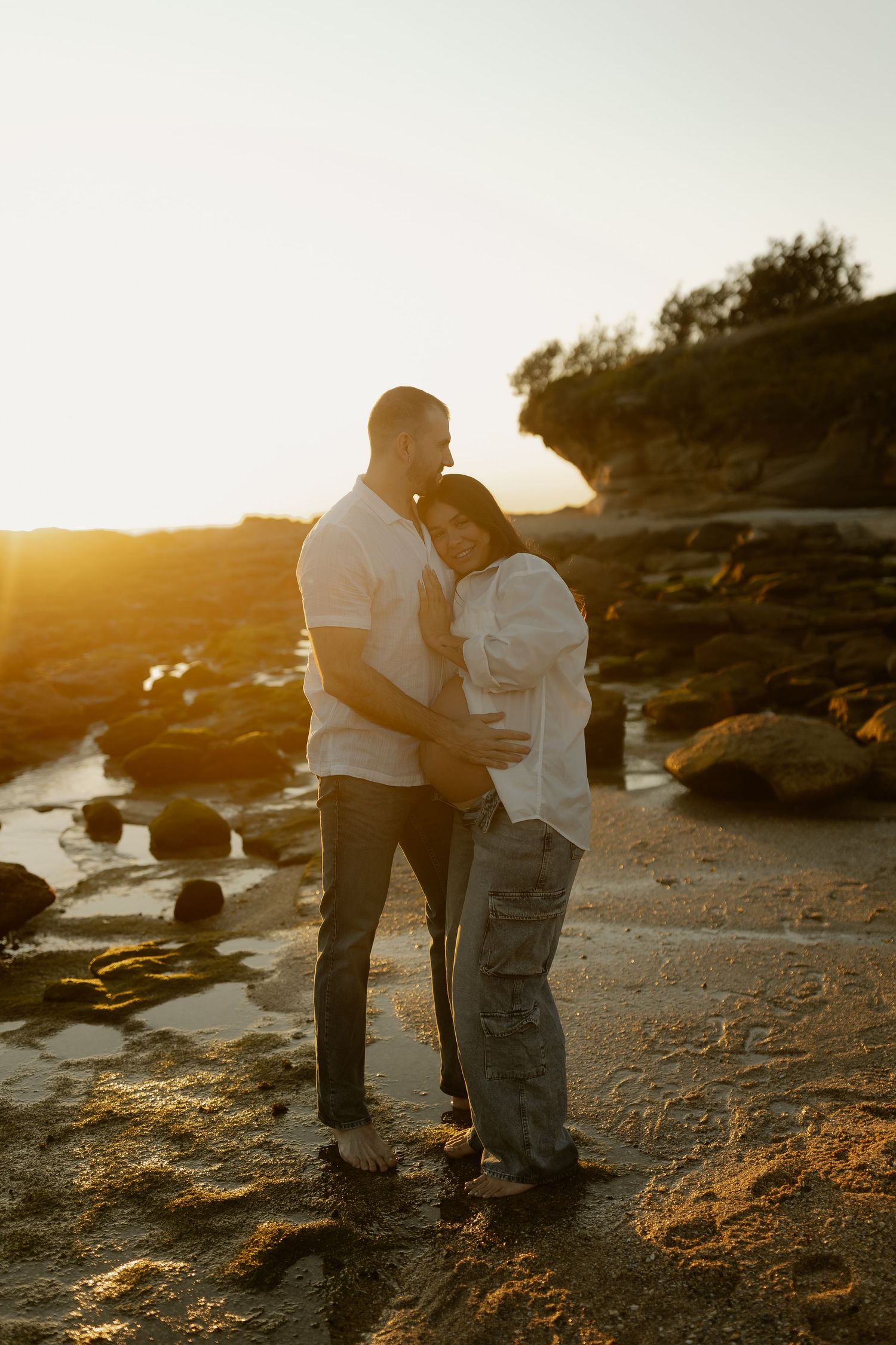 Couple embracing on rocky beach at golden sunset, pregnant woman cradling baby bump.