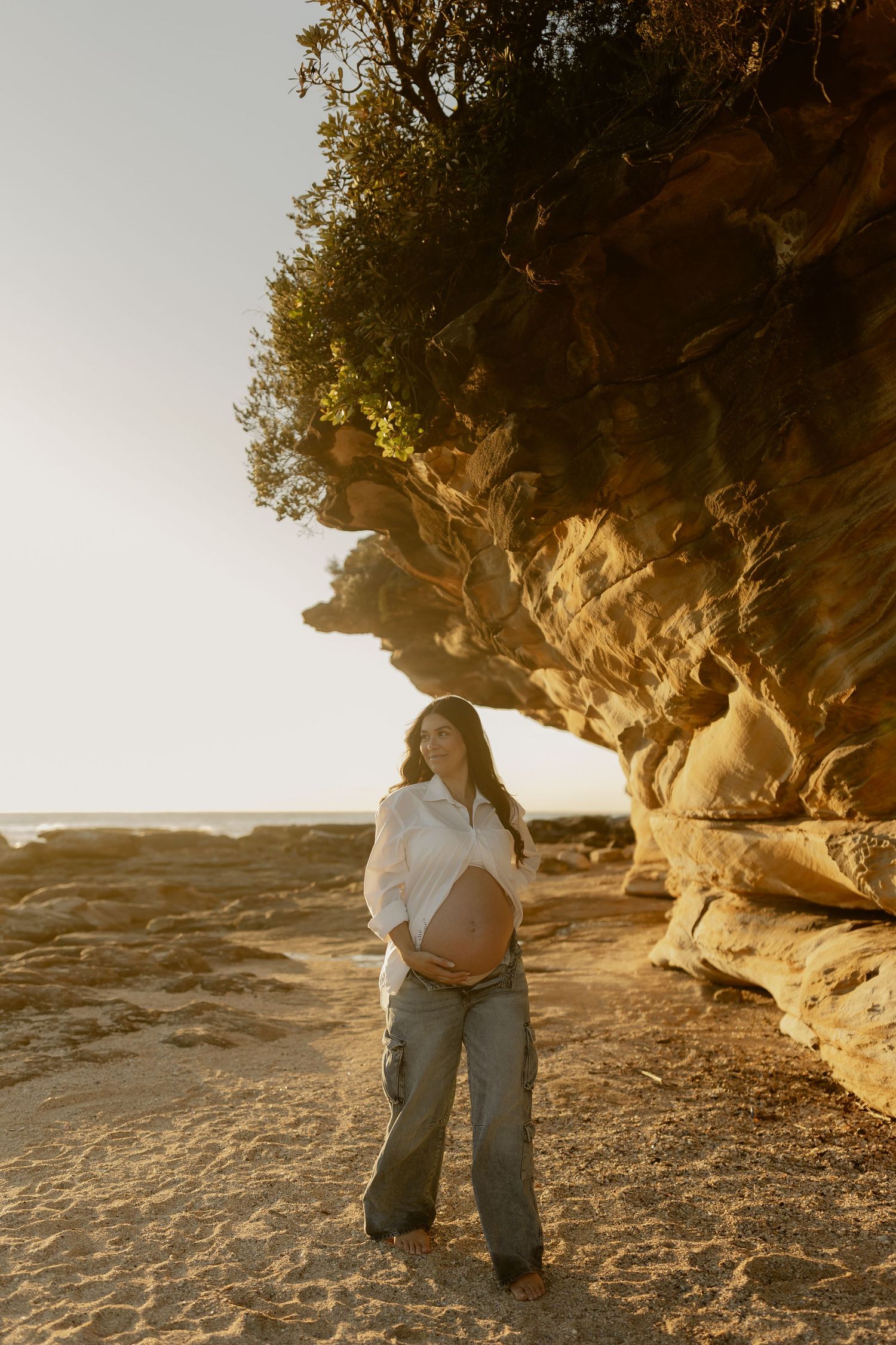 Pregnant woman in white shirt and jeans cradling baby bump beside golden sandstone cliffs at sunset beach.