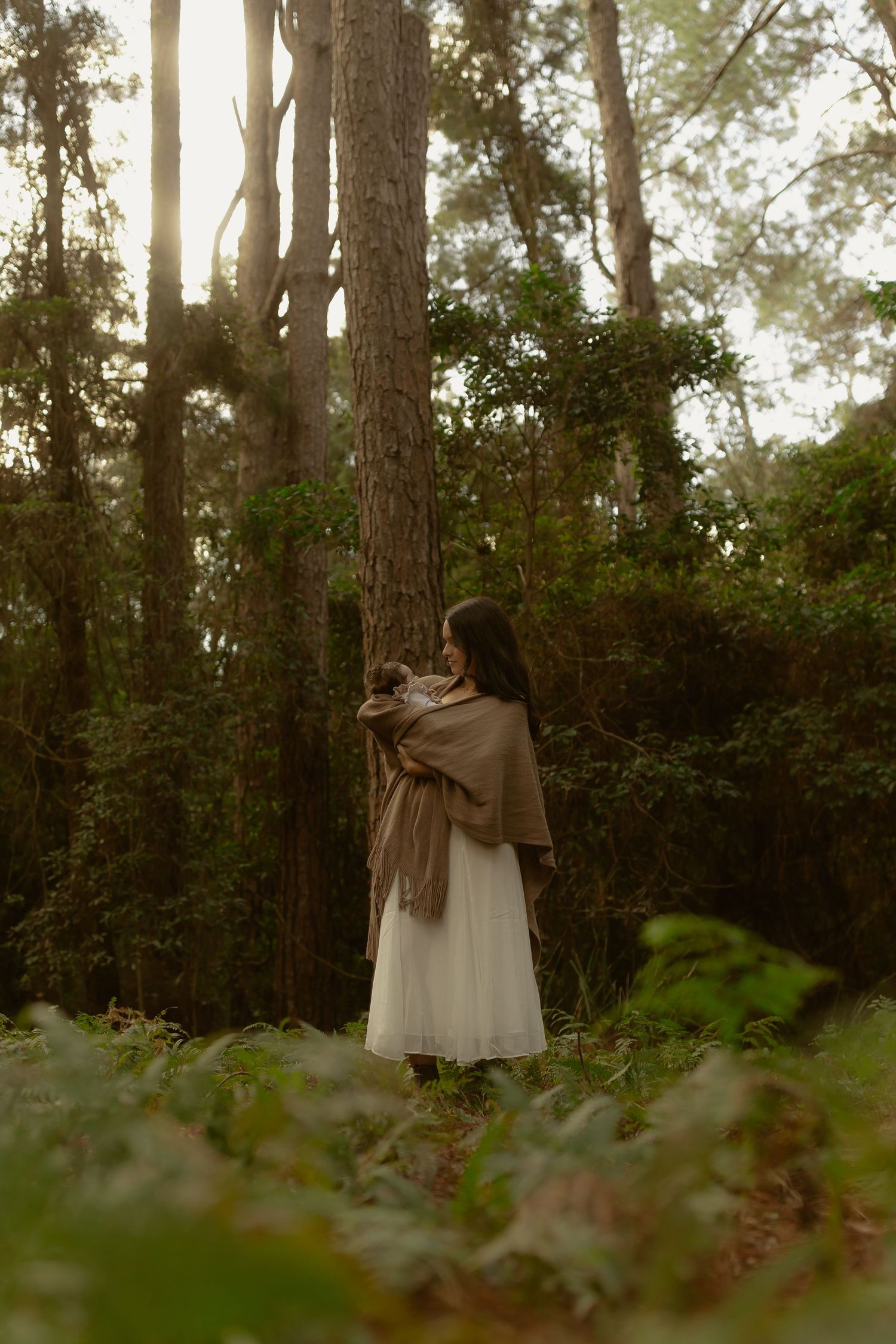 Woman in white dress and brown shawl holds baby beside tall pine tree in forest.