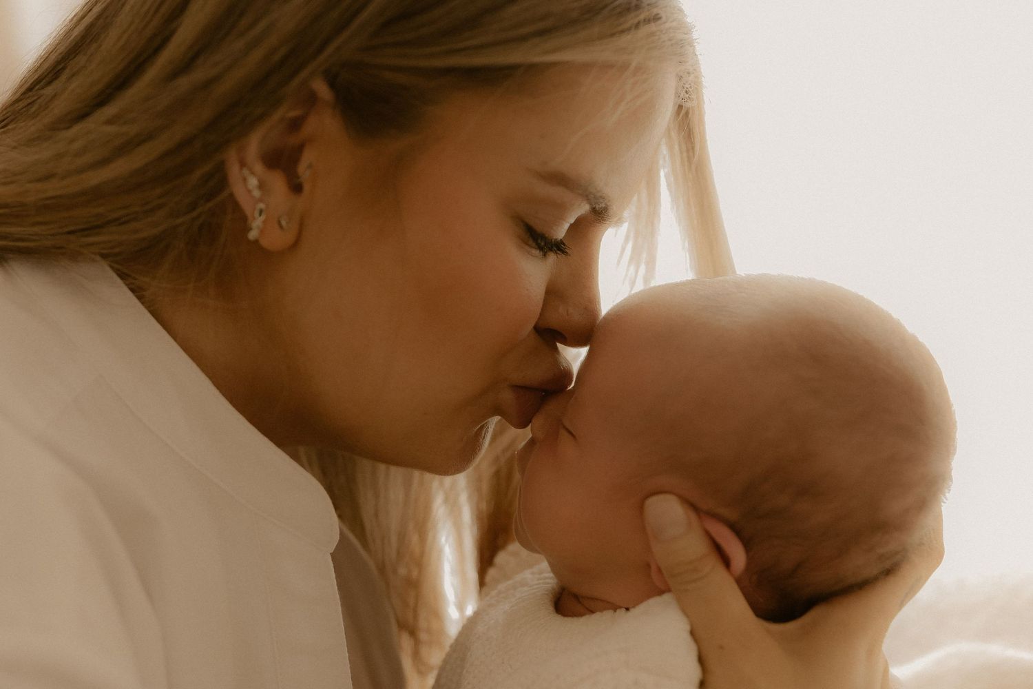 Tender moment captured as parent gives gentle kiss to baby's forehead in soft natural light.