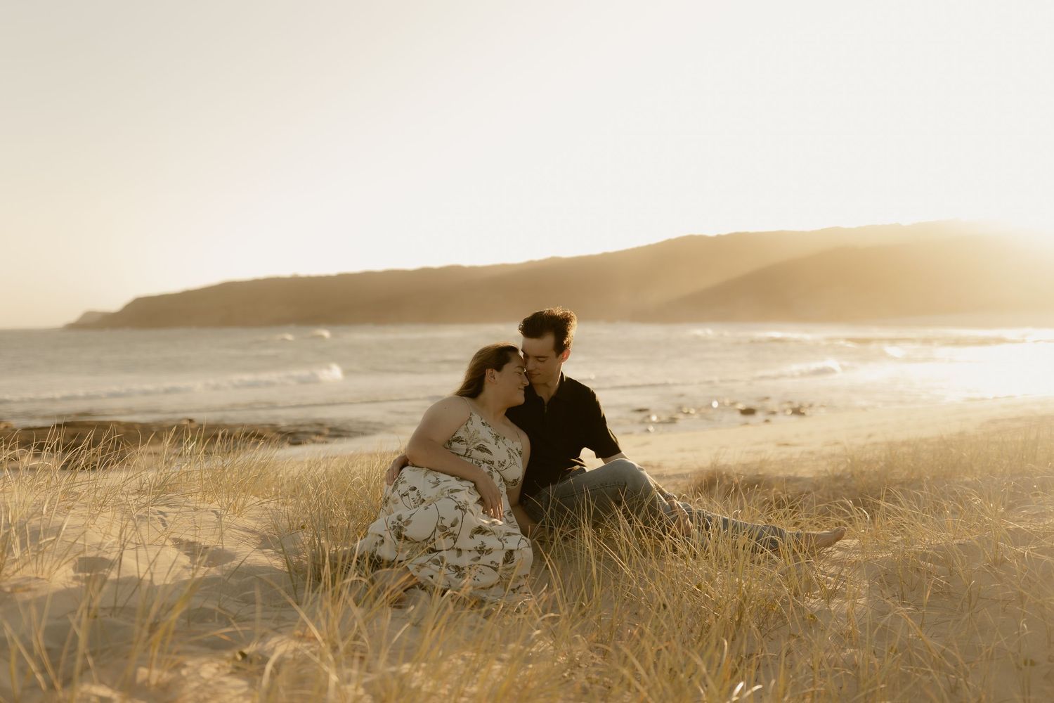 Silhouetted couple sits together on grassy coastal cliff overlooking ocean at golden sunset.