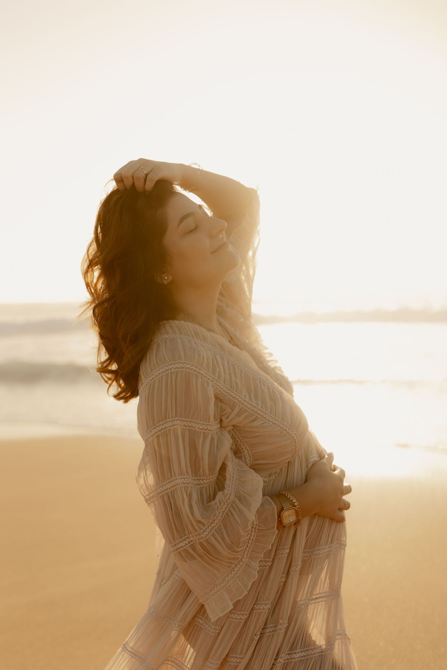 Dreamy backlit portrait of someone in white lace dress on sunlit beach at dusk.