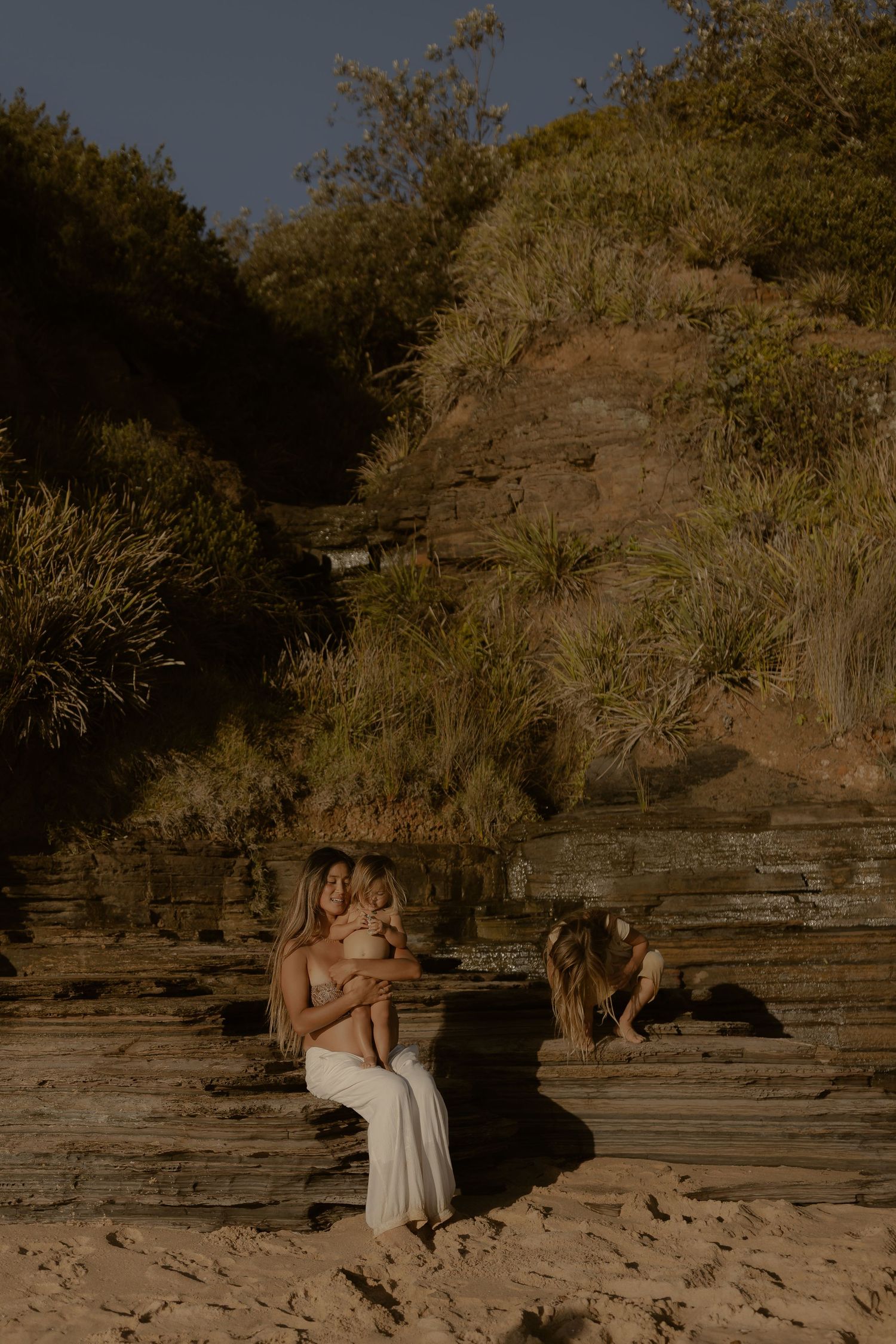 Intimate maternity photo session at rocky beach location with natural stone formations and warm lighting.