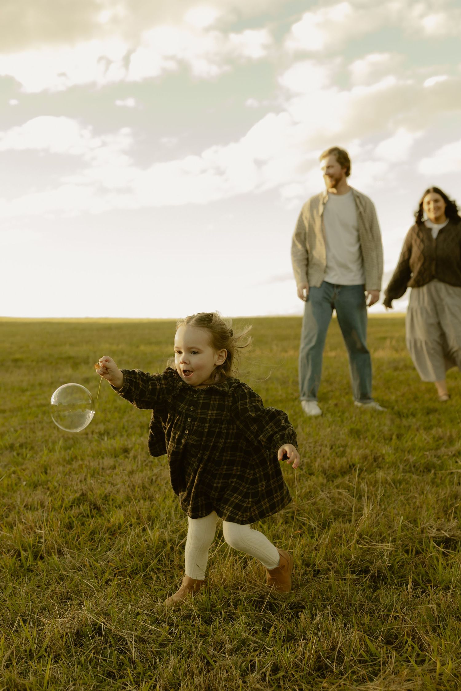 A toddler in a plaid dress chases a bubble in a green field while her parents watch in the background.