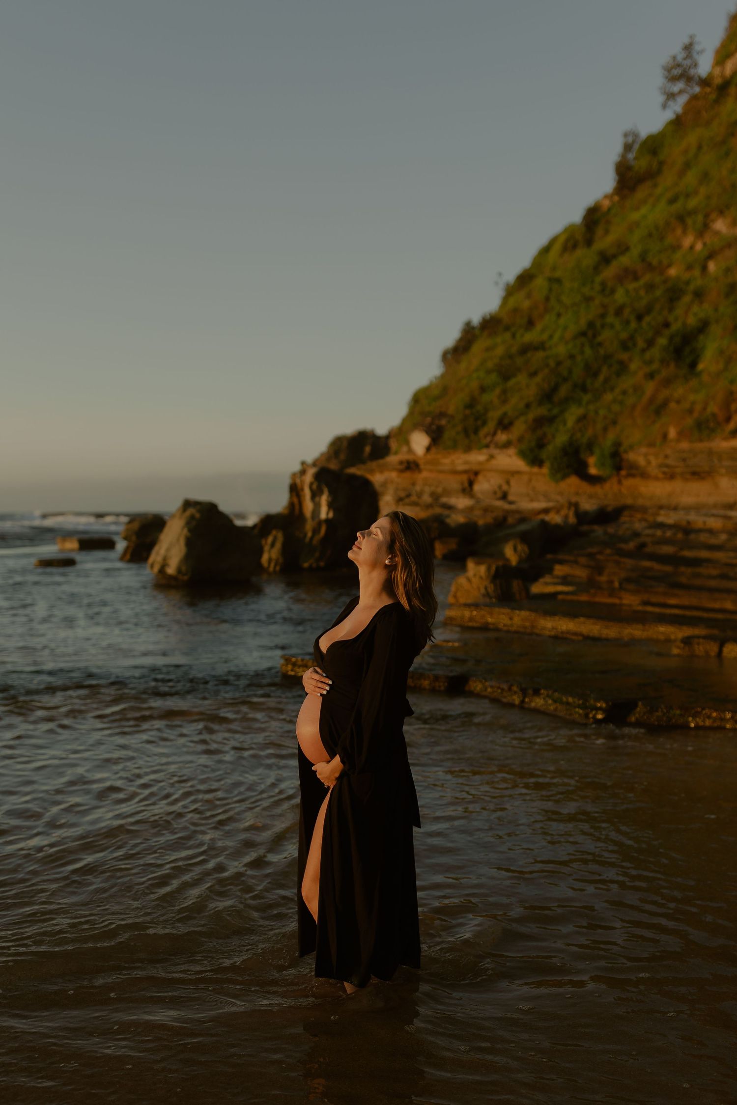 Pregnant woman in black dress standing in ocean water at golden hour near rocky coastal cliffs.