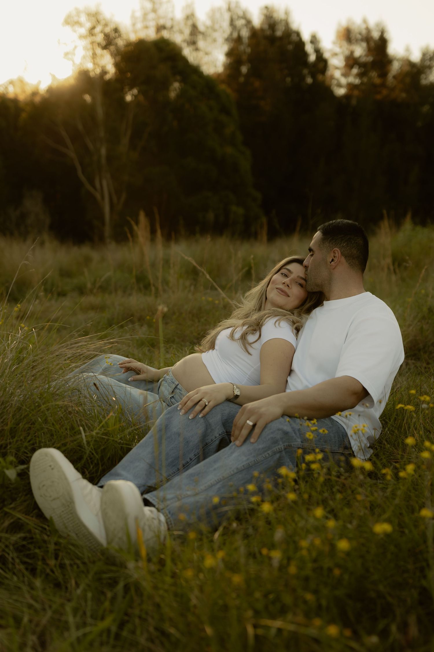 Pregnant couple relaxing together in a wildflower field during a warm sunset, sharing a tender moment.