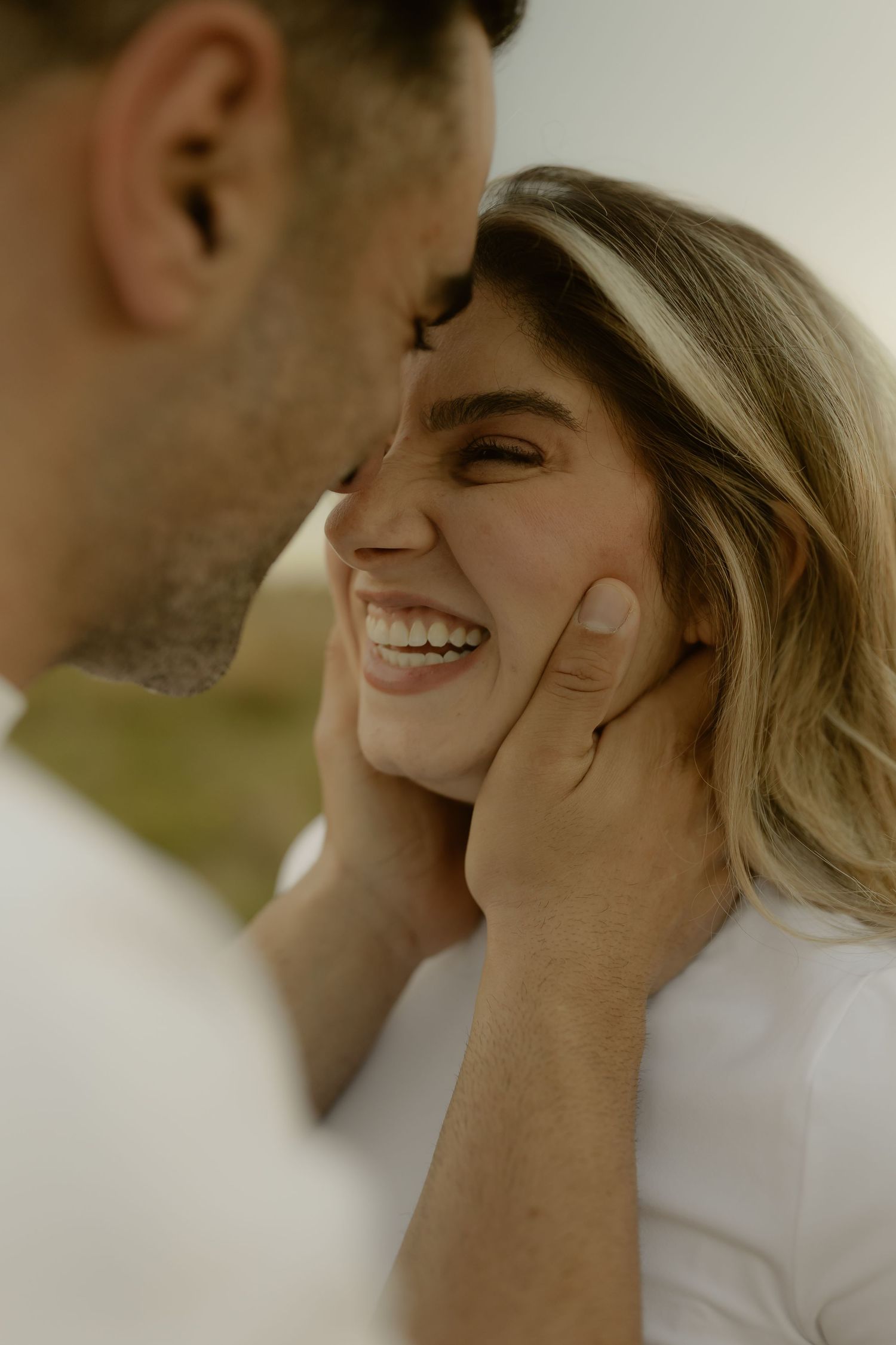 Man tenderly cupping smiling womans face while leaning in for a kiss outdoors.