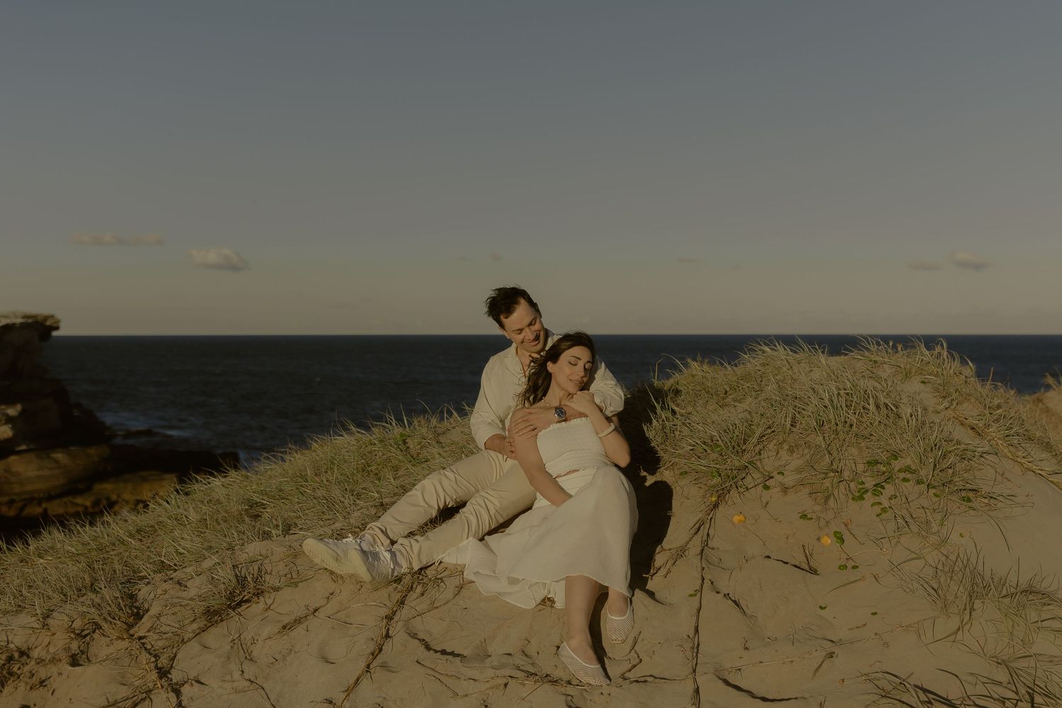 Couple sitting together on sandy coastal dunes at sunset, with ocean and clear sky in the background.