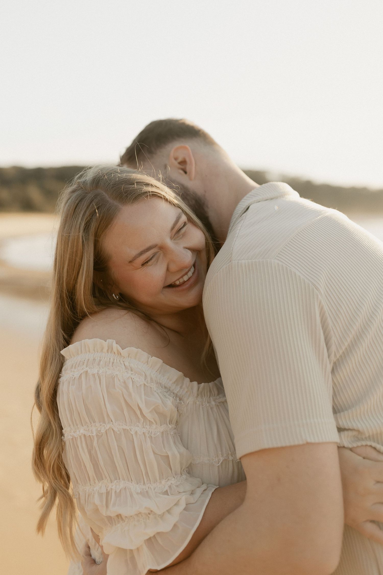 Couple embracing on a sunny beach, woman smiling joyfully in white off-shoulder dress.