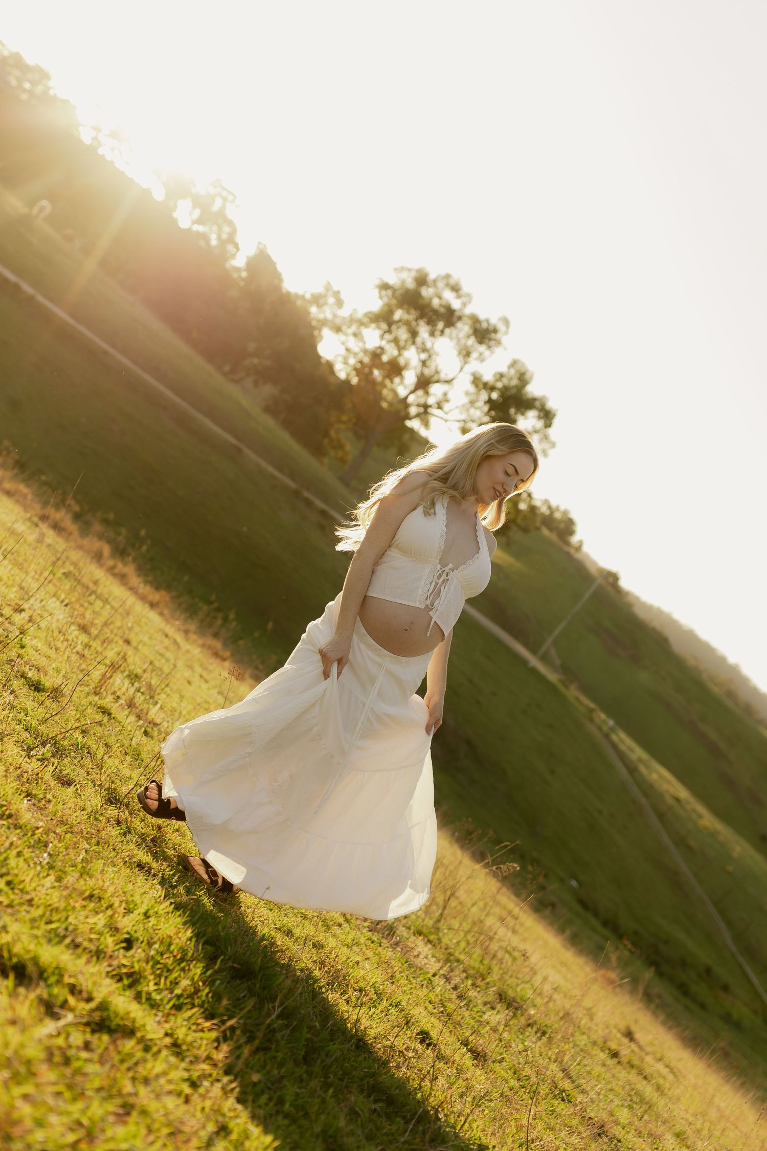 Woman in white crop top and flowing skirt walking through sunlit green field at golden hour.