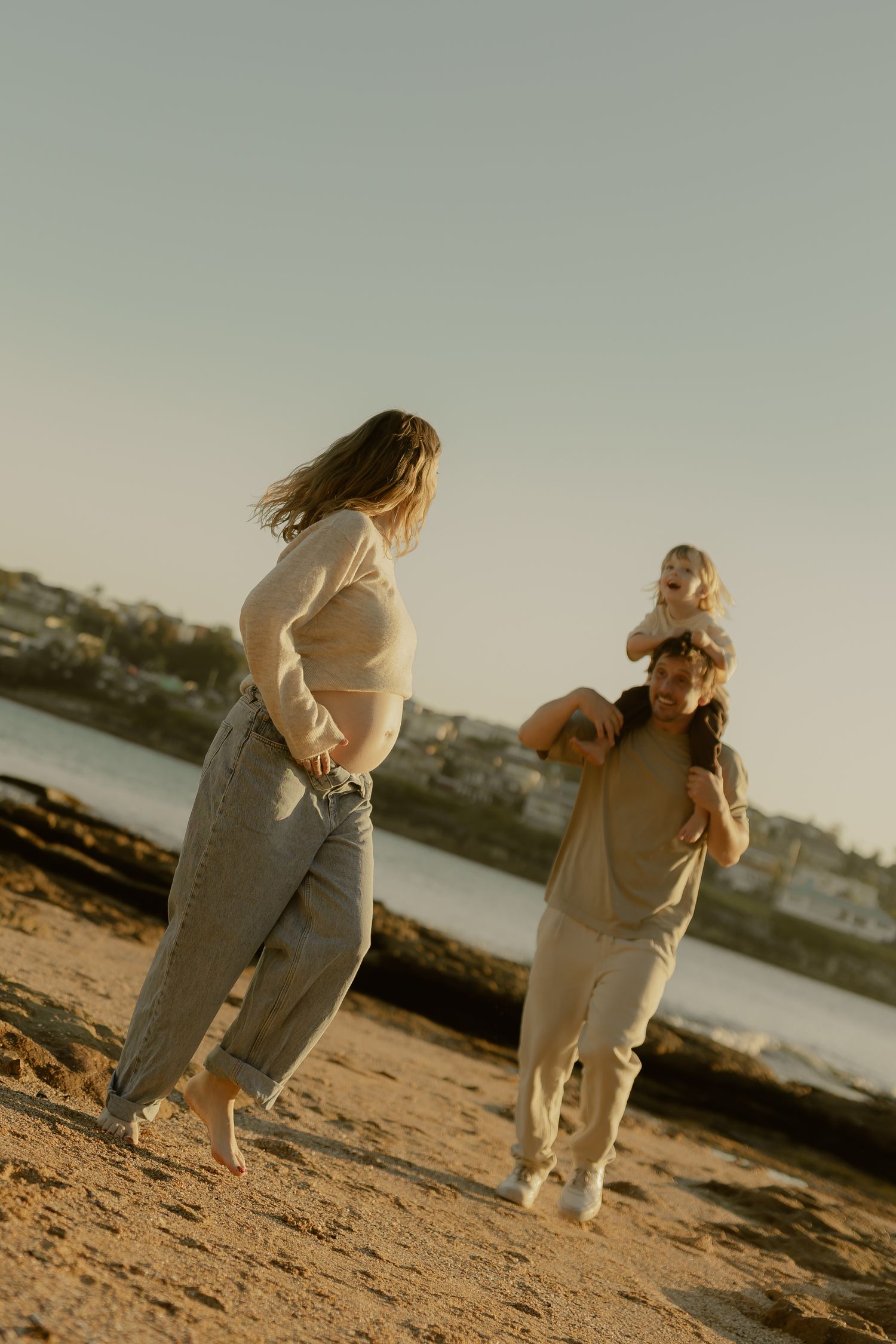 Pregnant woman and partner with toddler on shoulders walking on sandy beach at sunset.