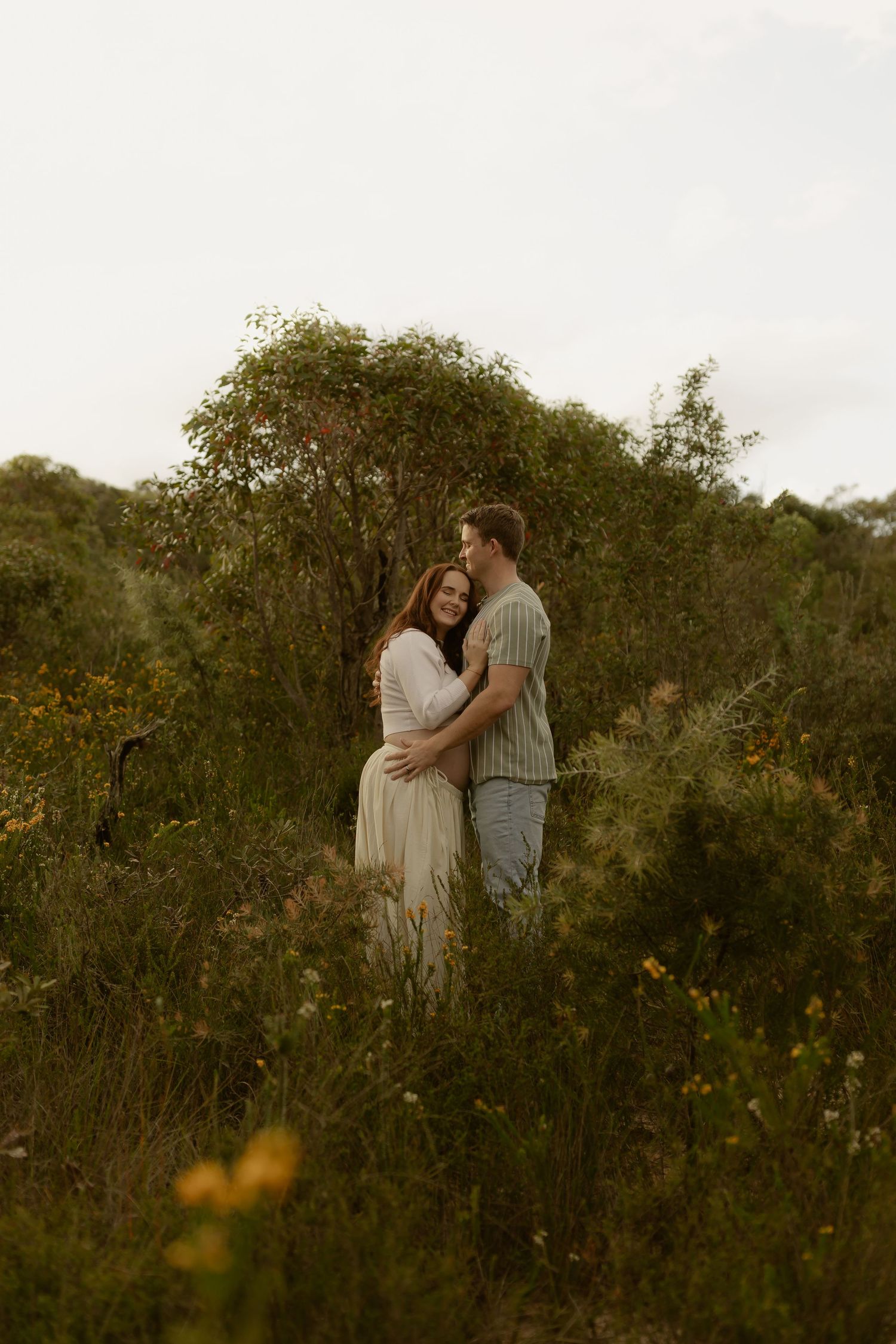 Couple embracing in lush green field with wildflowers, woman in white dress smiling, romantic outdoor portrait.
