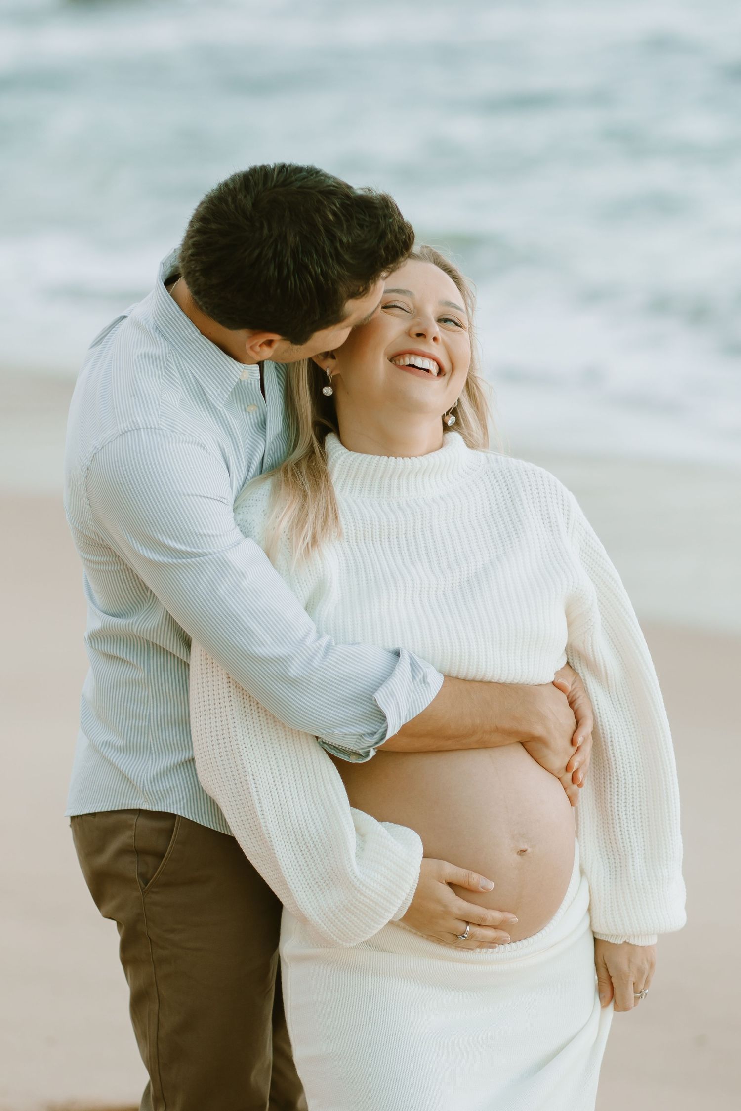 A joyful pregnancy photoshoot on the beach features a couple in white sweaters embracing the baby bump.