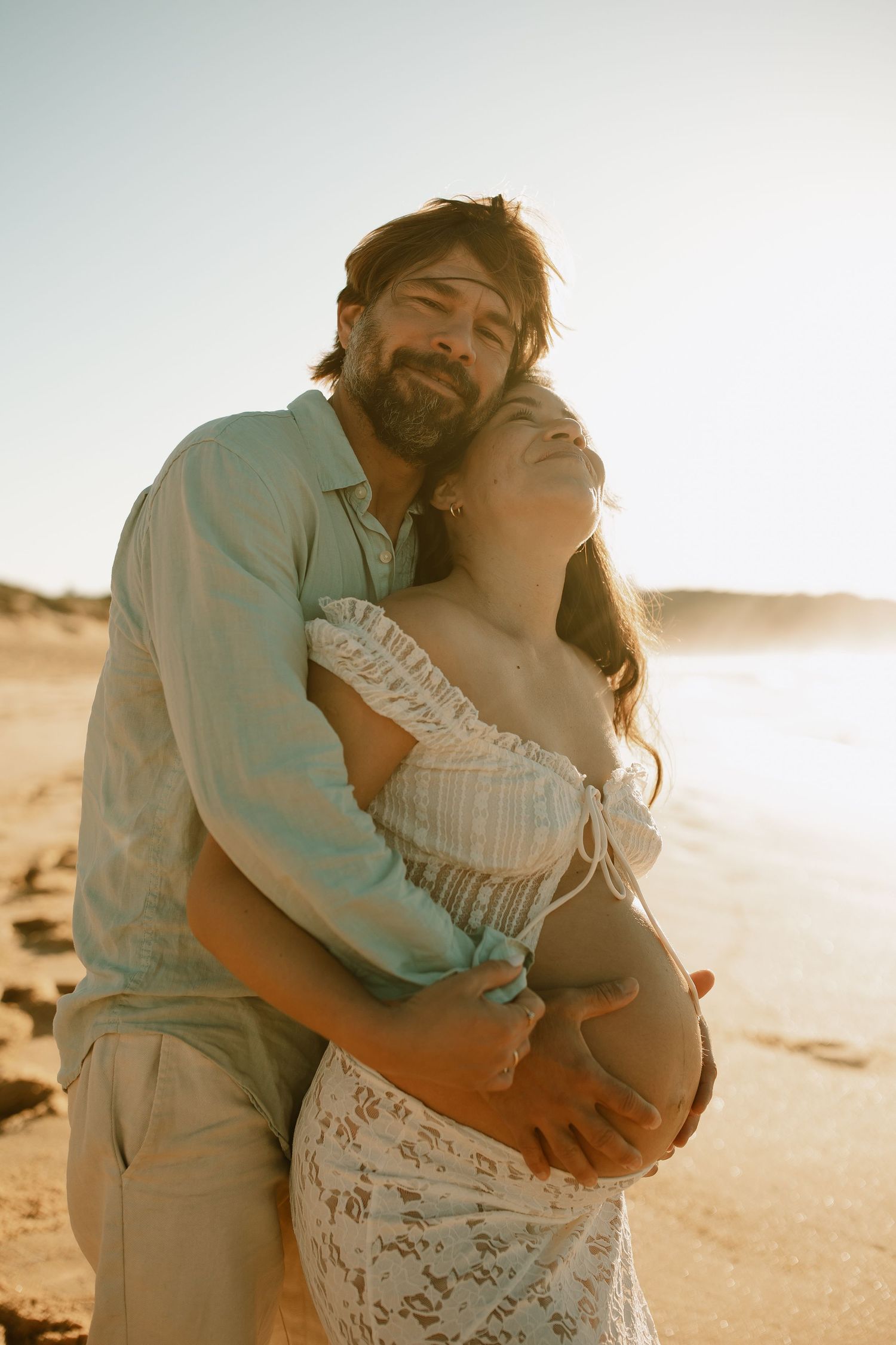 Expecting couple embraces tenderly on beach at sunset in romantic maternity photoshoot.