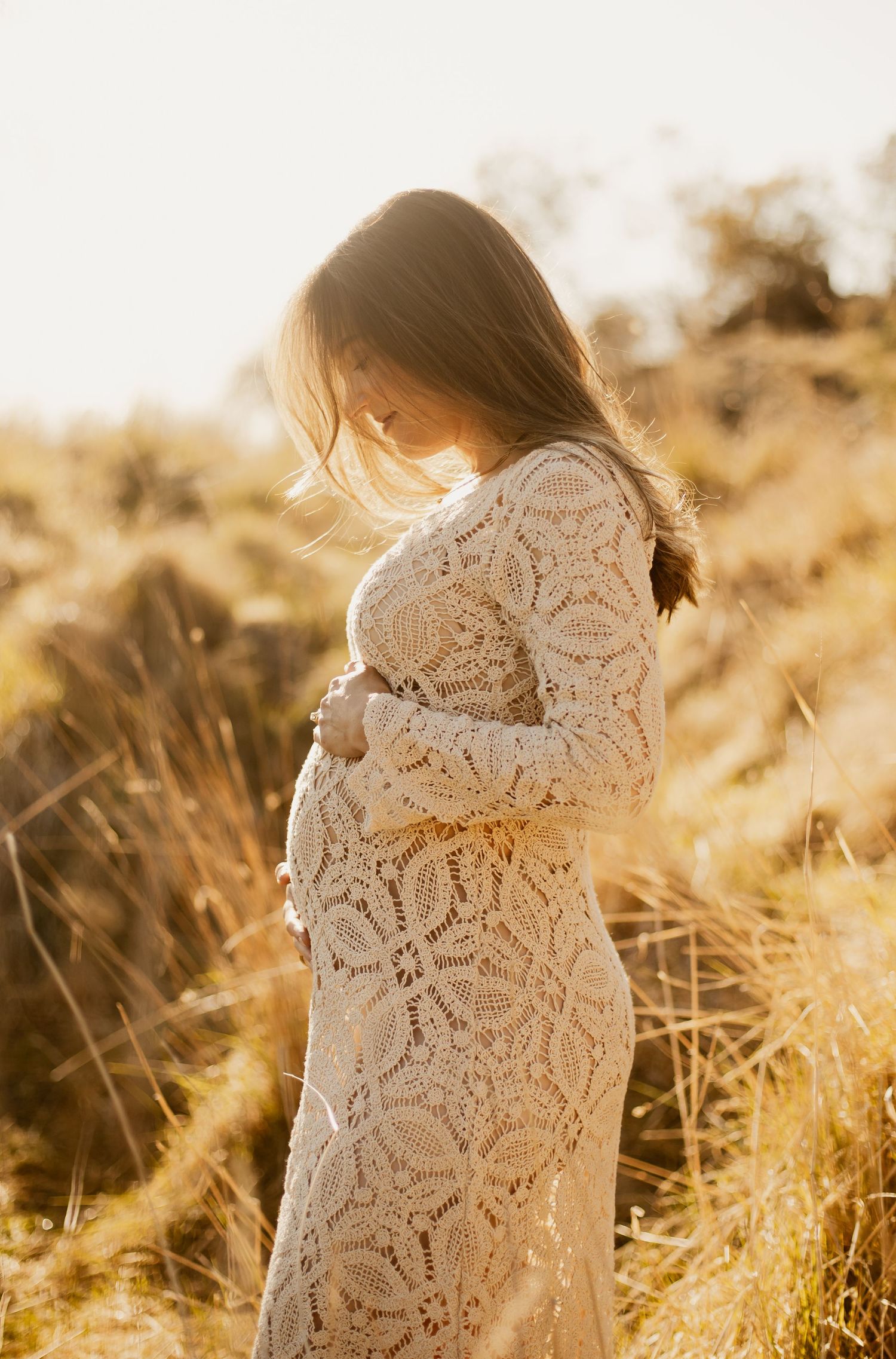 Silhouette of a woman in a lace white dress standing in golden grass during sunset.
