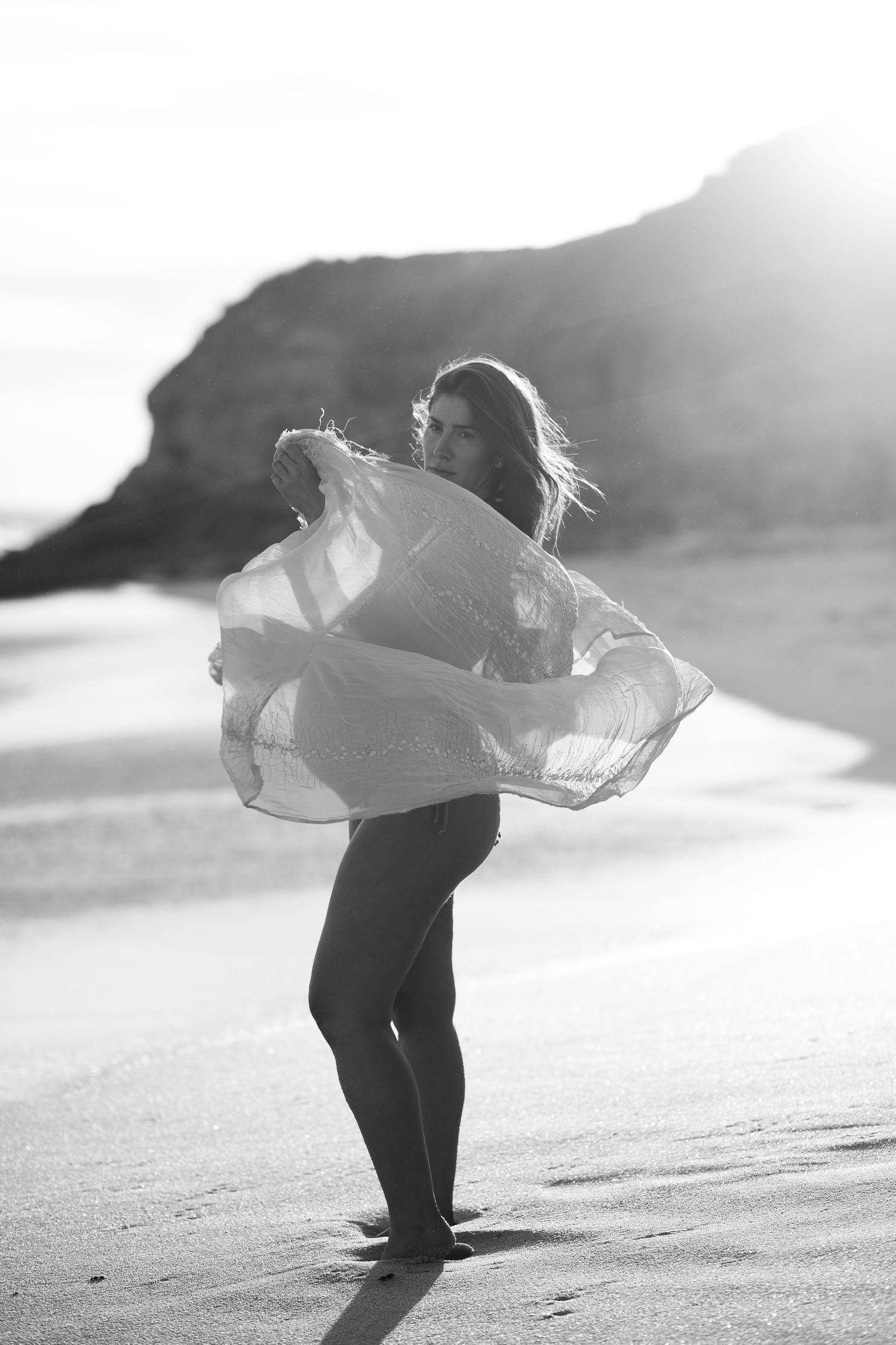 A silhouetted figure dances with flowing white fabric on a beach at sunset in a dreamy black and white photograph.