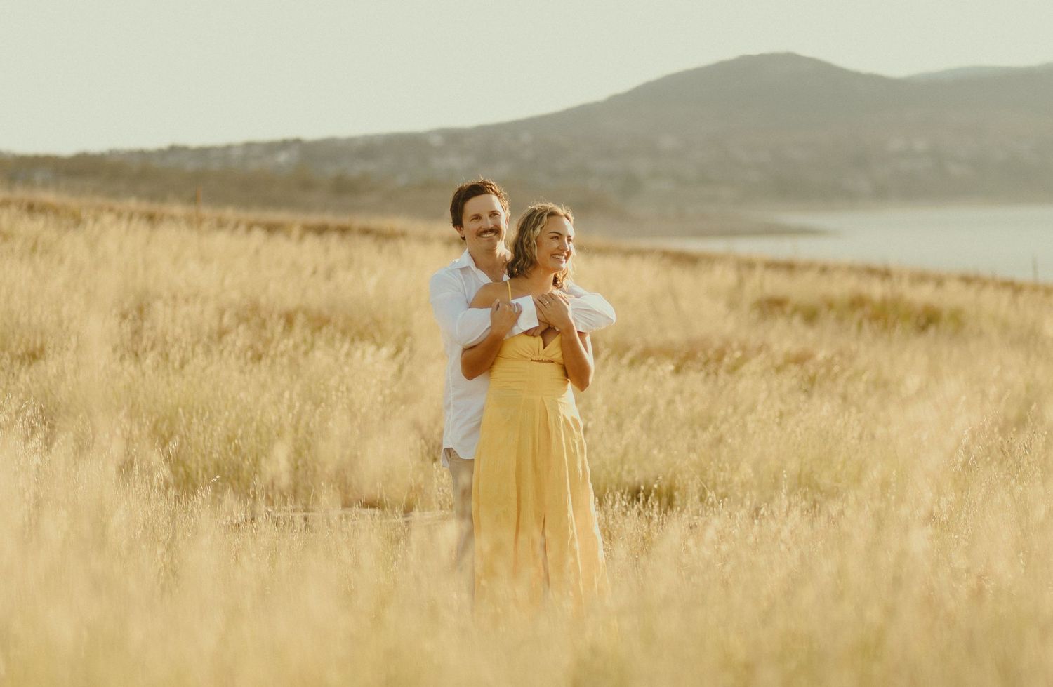 A couple embraces in a golden wheat field at sunset with rolling hills and a lake in the background.