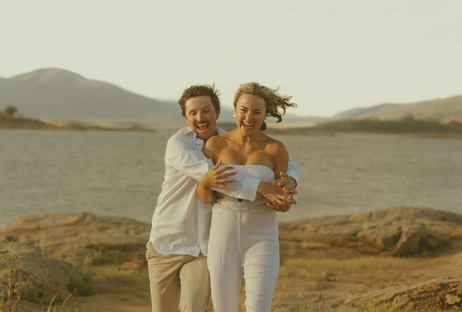 A couple embraces while laughing on a hilltop during golden hour with mountains in the background.