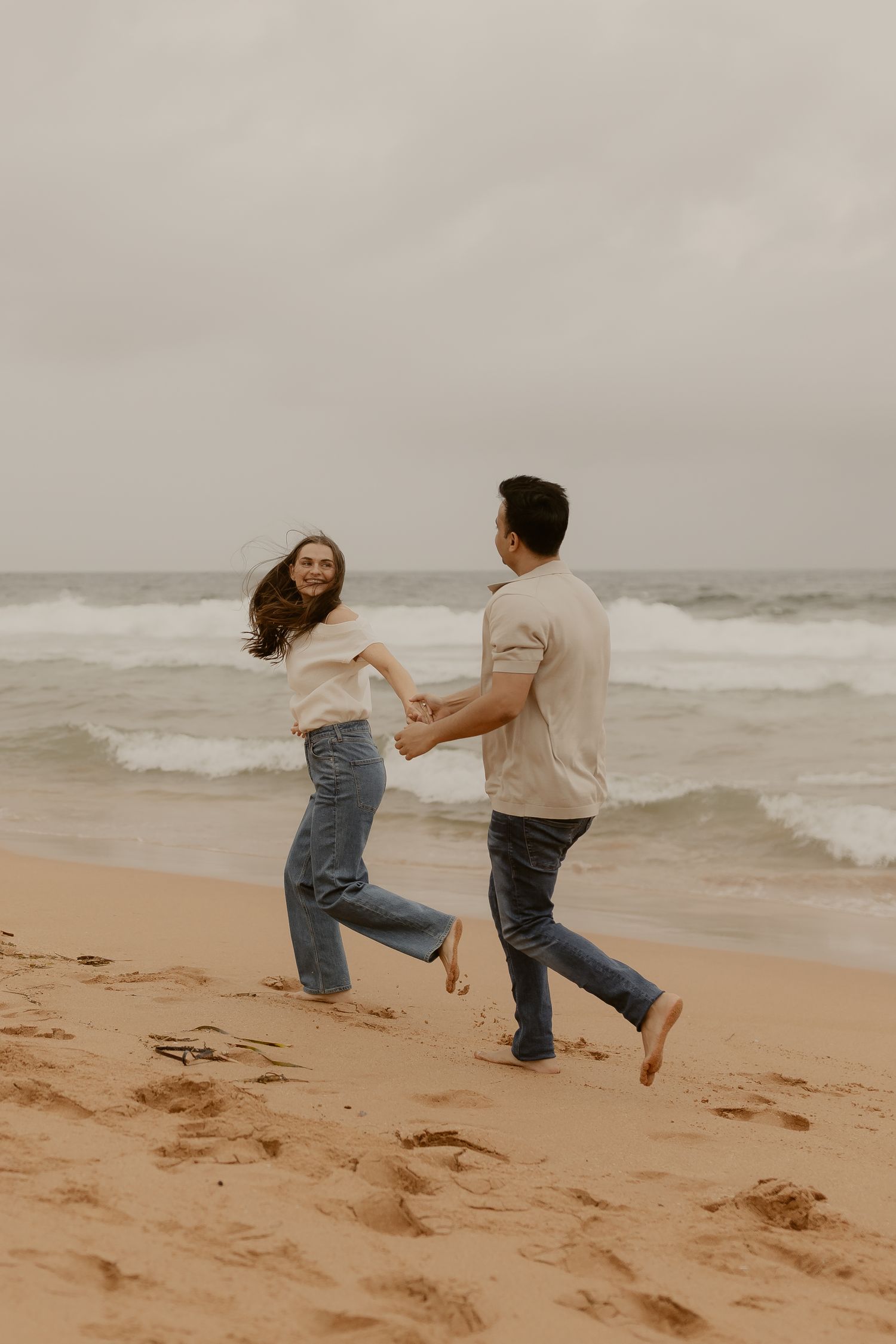 A playful couple runs and dances together on a sandy beach with crashing waves in the background.